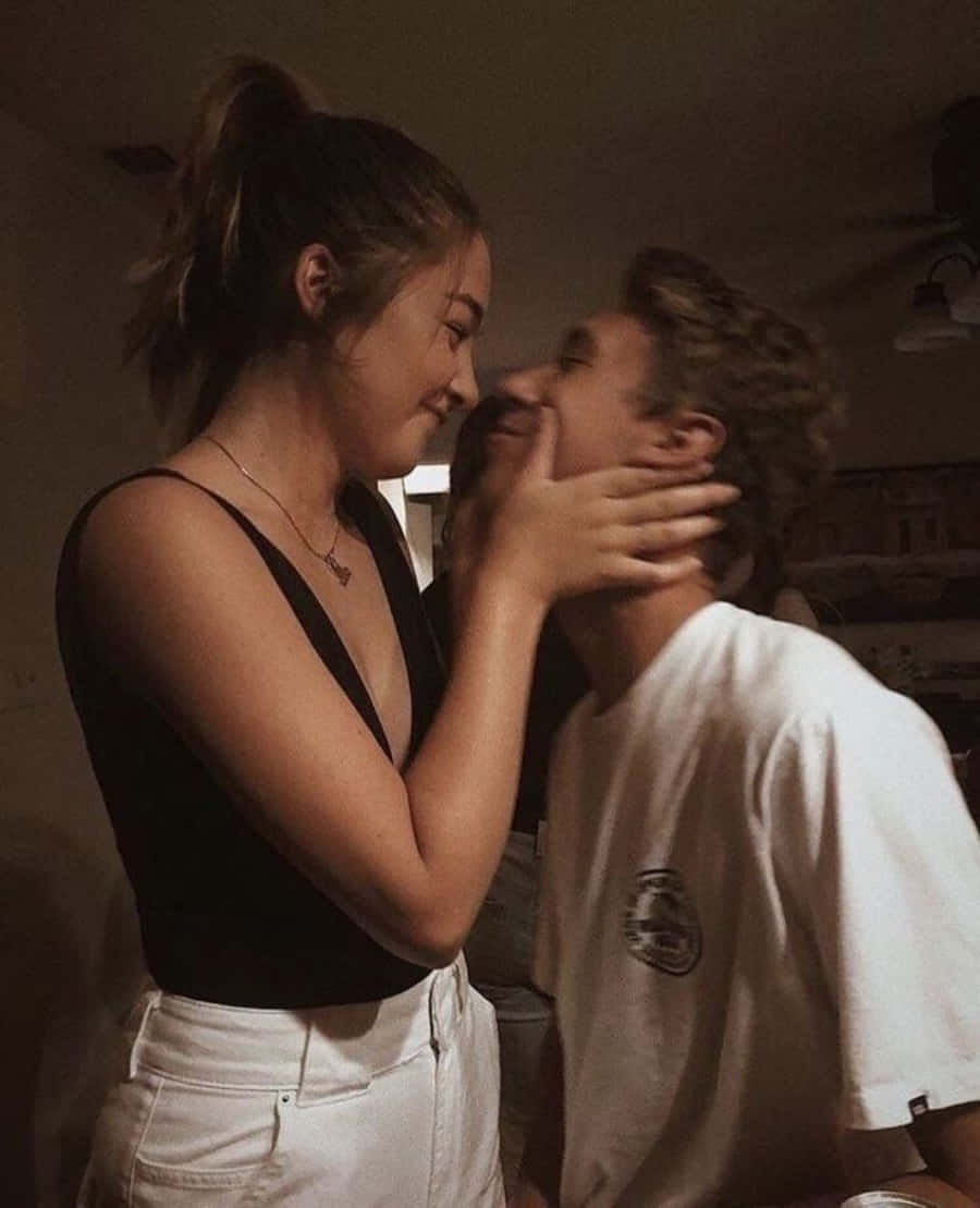 Aesthetic Couple 桌布 壁紙 — 一個溫暖燈光的室內場景捕捉到一個溫柔的時刻，一位女士用手輕輕托著男士的臉龐，兩人互相微笑。