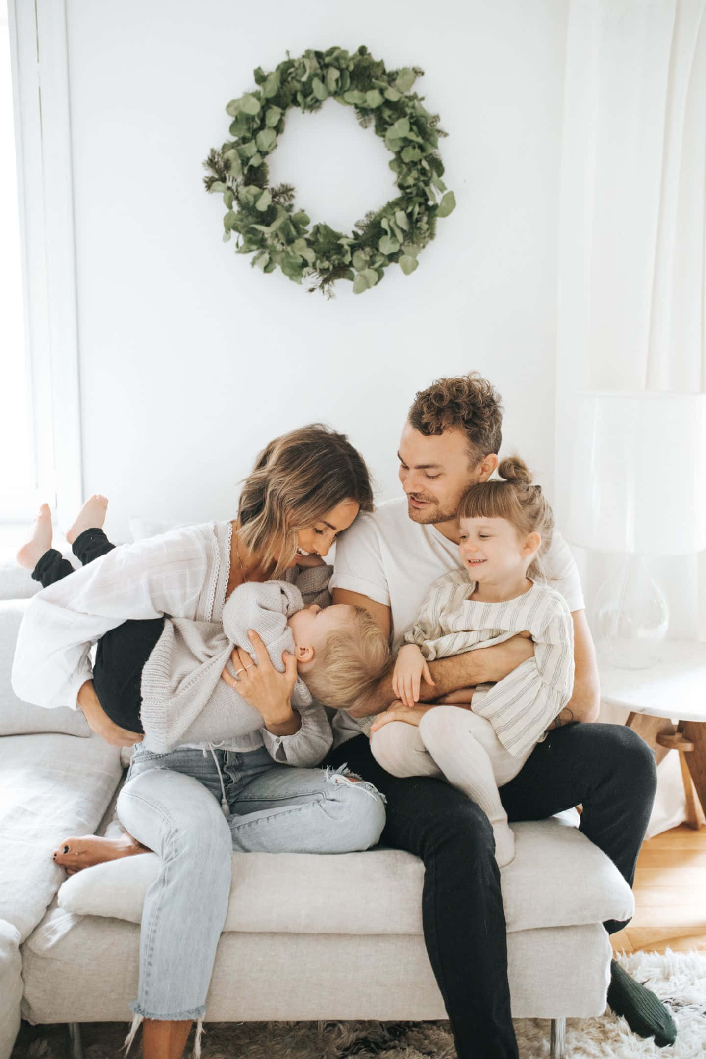 Family Christmas 桌布 壁紙 — 一個溫暖的四口之家在淺色沙發上依偎在一起，身後的白牆上掛著一個綠色花環。