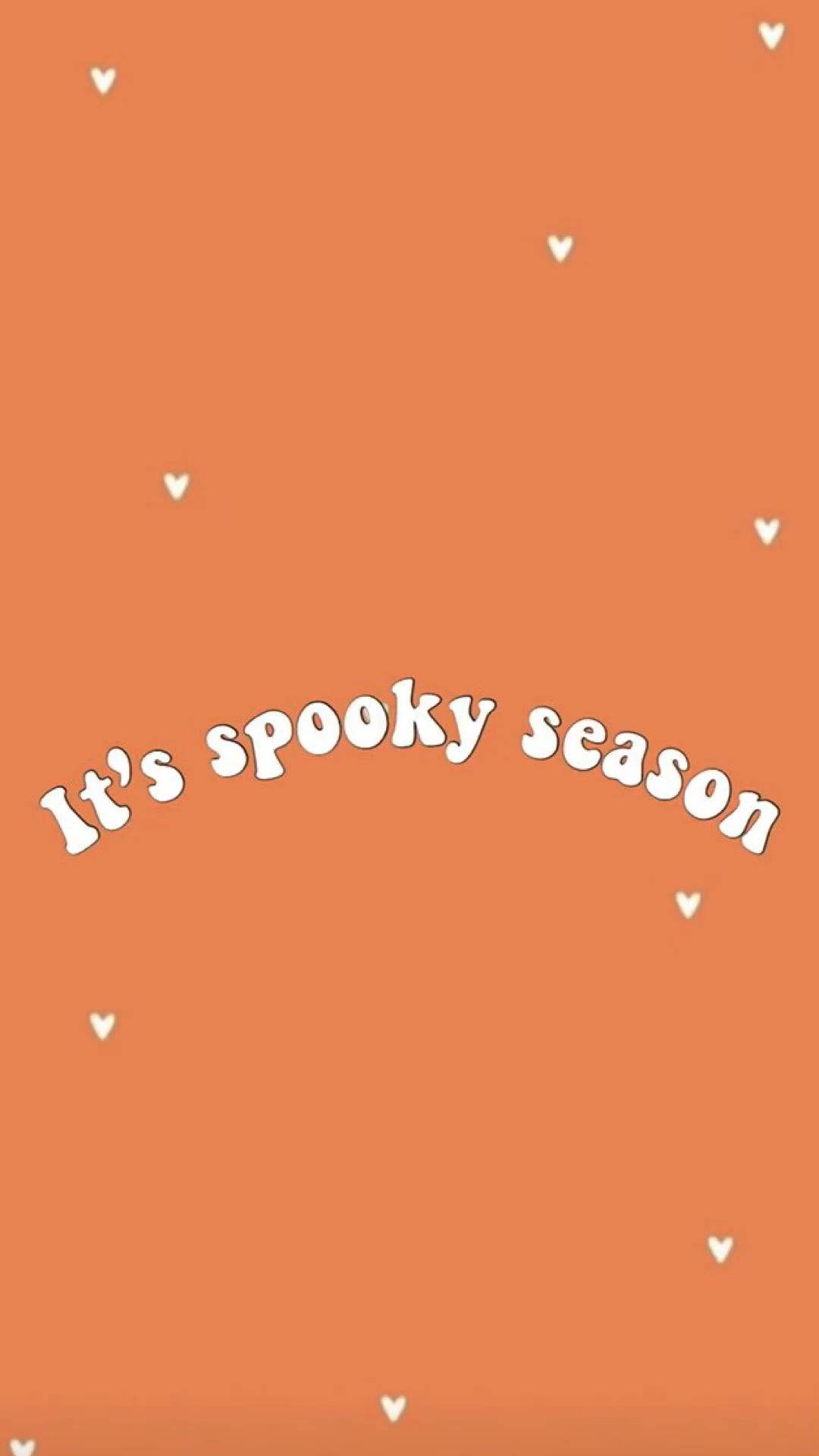 Spooky Aesthetic 桌布 壁紙 — 一個溫暖的橙色背景，散落著白色心形圖案，並以俏皮的白色字體寫著「It's spooky season」。