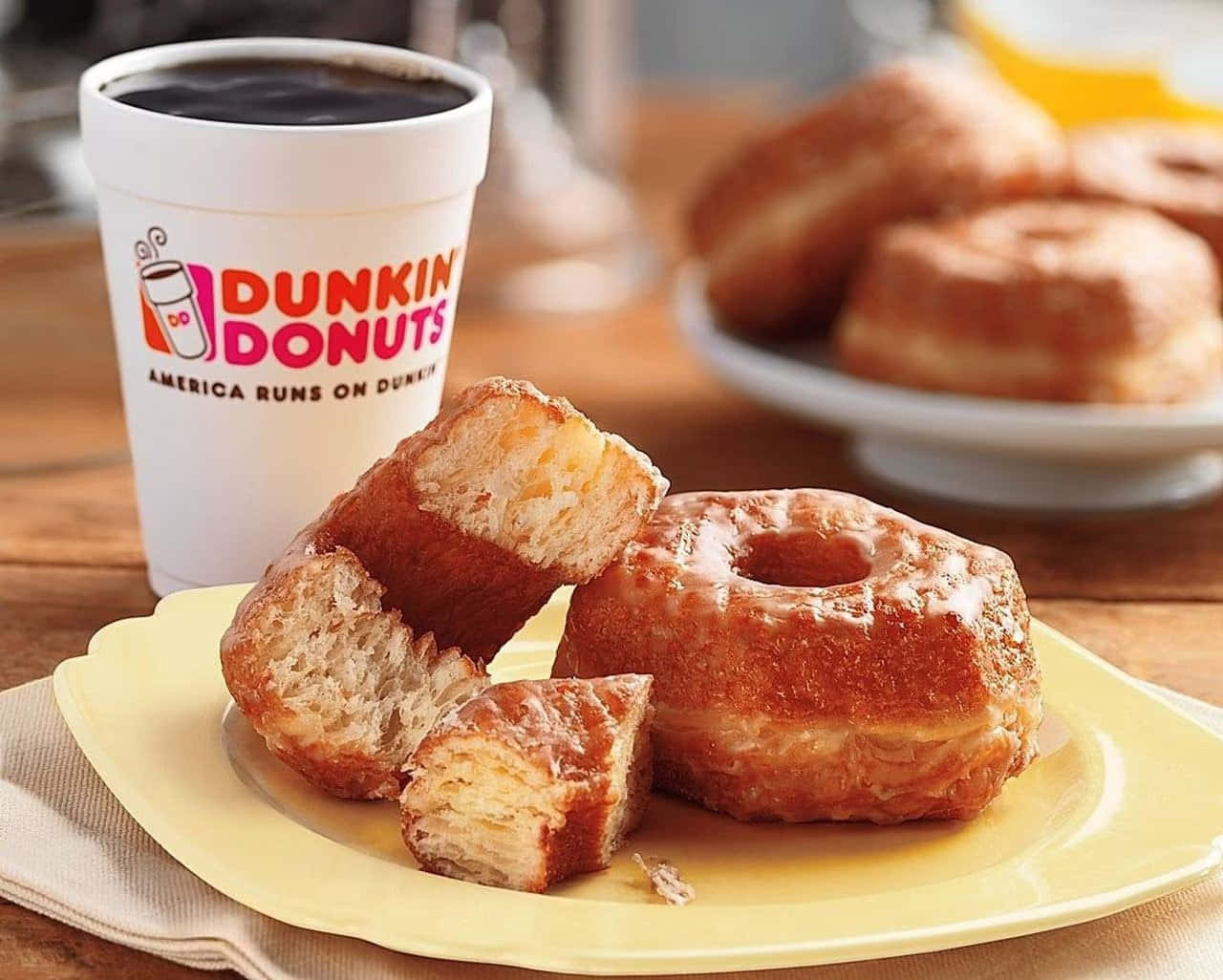 Dunkin Donuts 桌布 壁紙 — 一個溫暖的糖霜甜甜圈被掰開一半，放在一個黃色盤子上，旁邊有一杯Dunkin' Donuts咖啡。