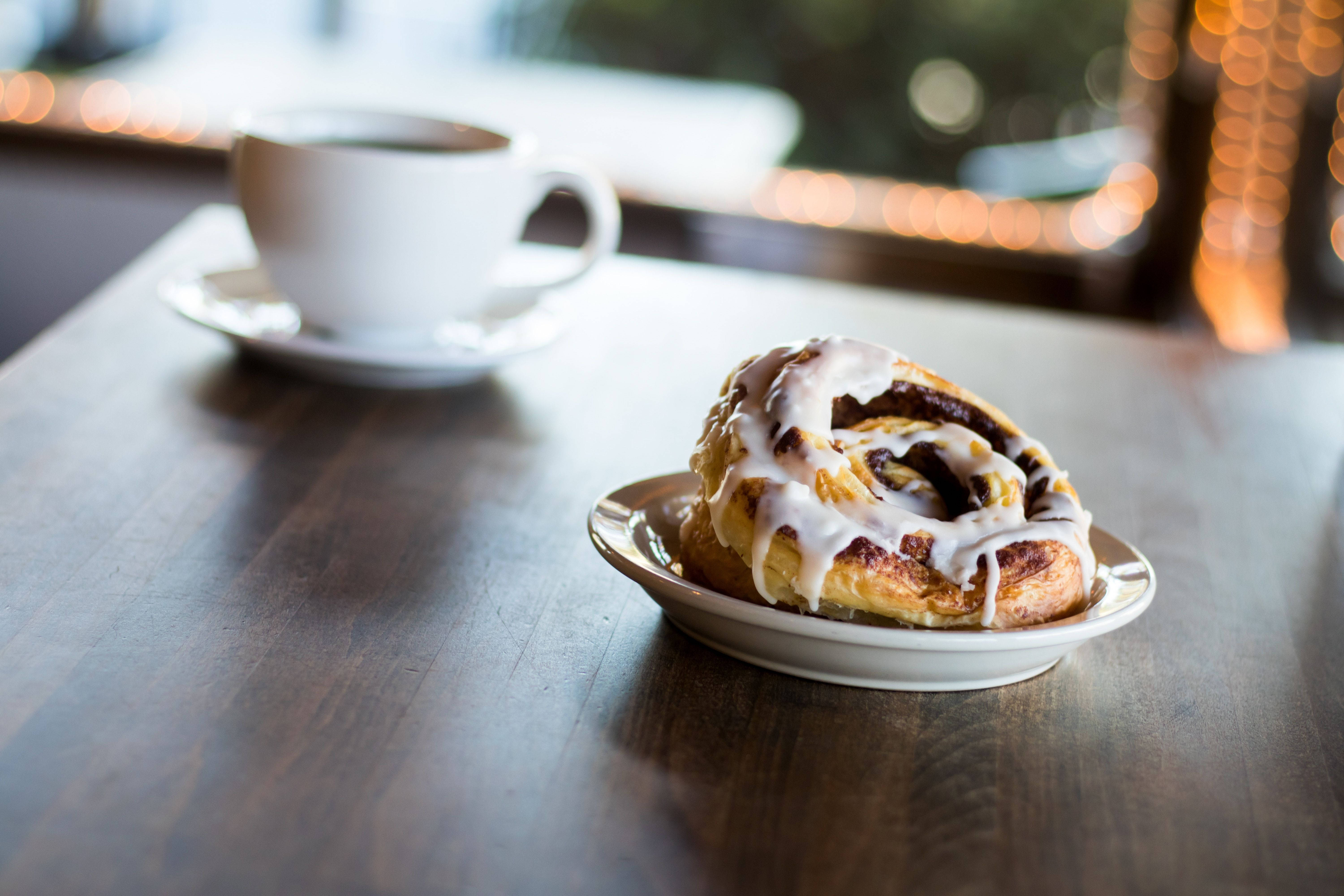 Pastry 桌布 壁紙 — 一個溫暖的肉桂捲配白色糖霜，旁邊放著一杯咖啡，放在木桌上，背景有散景燈光。