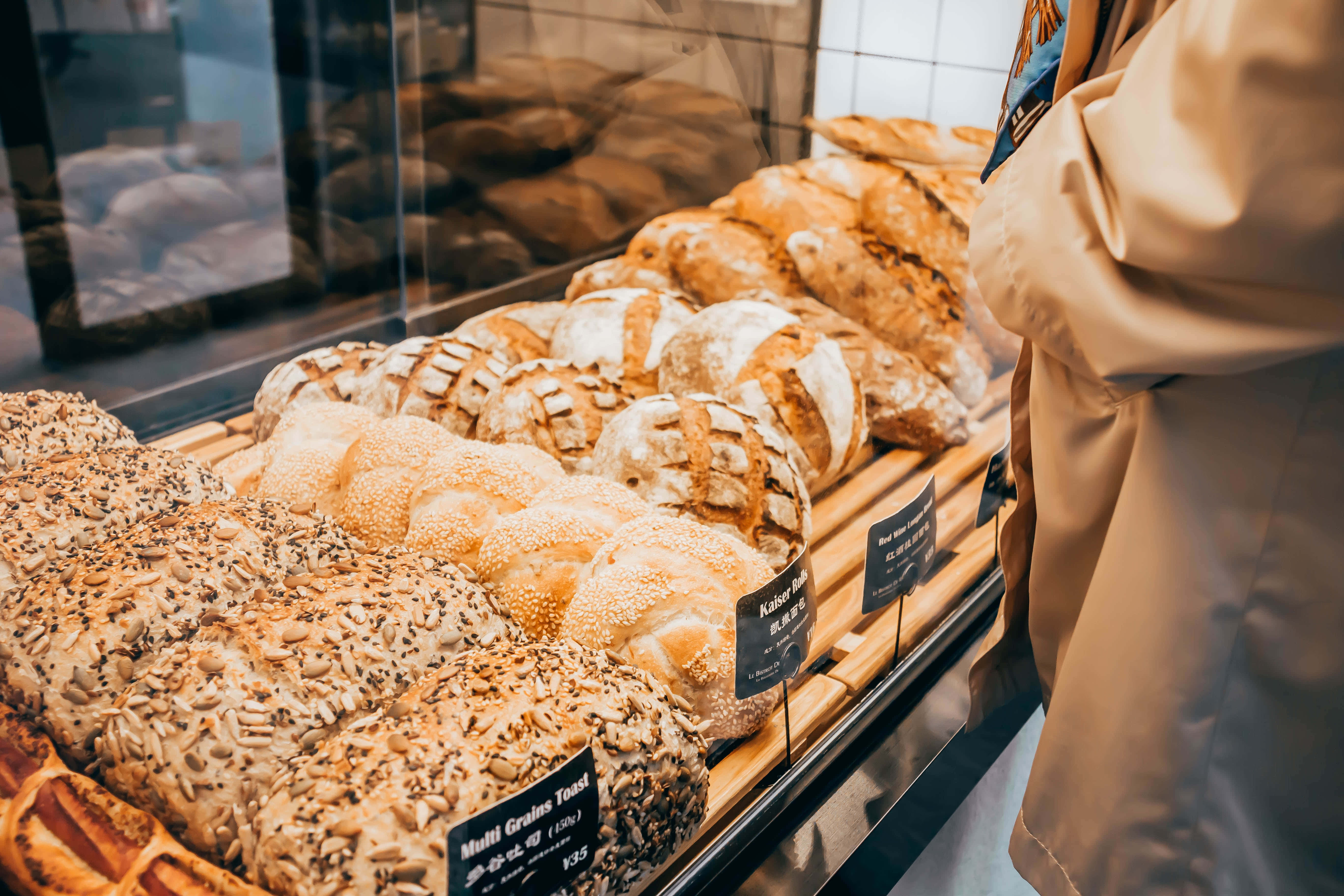 Bakery 桌布 壁紙 — 一個溫暖誘人的麵包店展示櫃，裡面裝滿了金黃酥脆的麵包和種子麵包，有一個穿著米色外套的人在瀏覽。