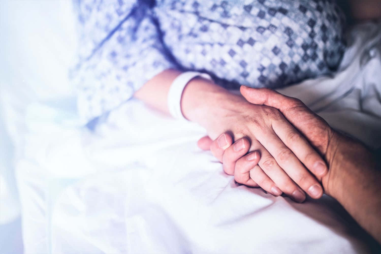 Holding Hands 桌布 壁紙 — 一個溫柔的時刻，兩隻手在醫院病床上緊握在一起，一隻戴著手環，另一隻輕輕地放在上面。