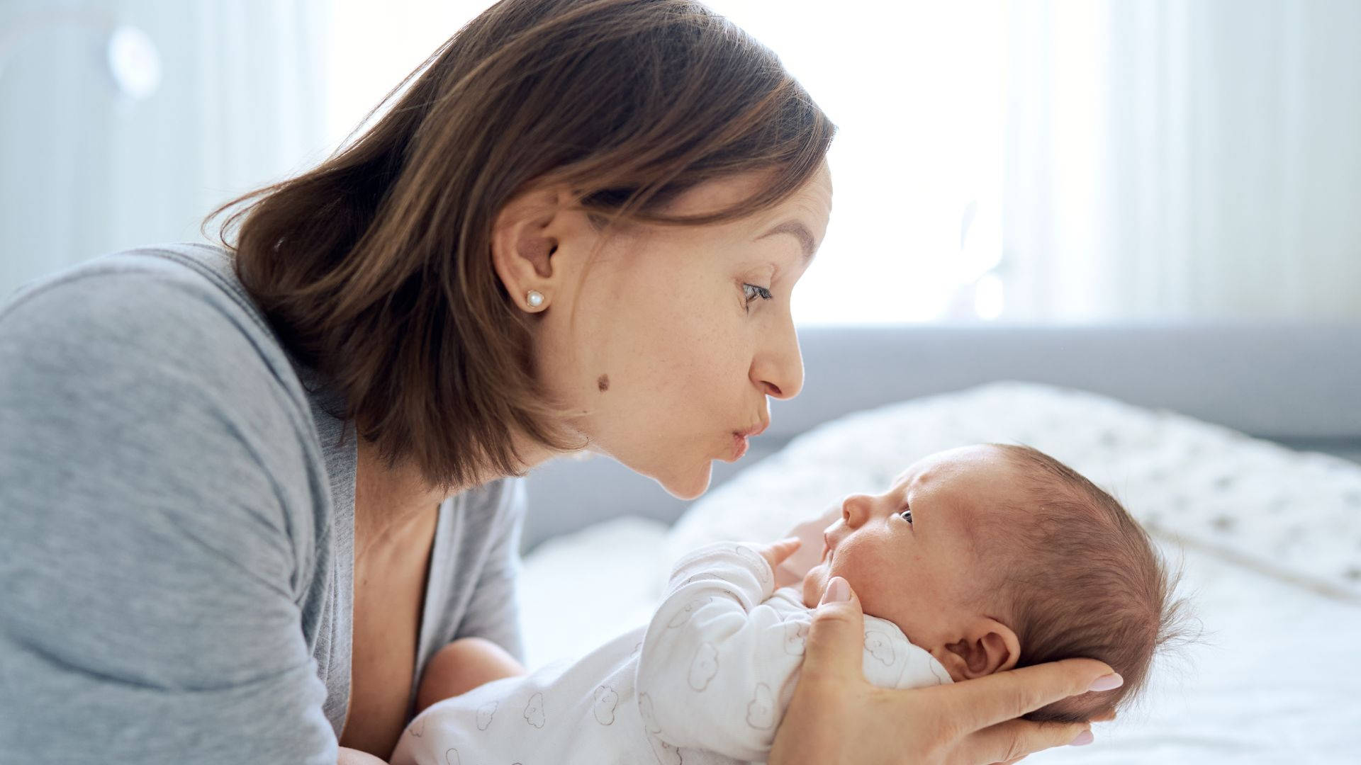 Mom 桌布 壁紙 — 一個溫柔的時刻，母親在柔和的燈光下輕輕親吻新生嬰兒的額頭，緊緊抱著他們。