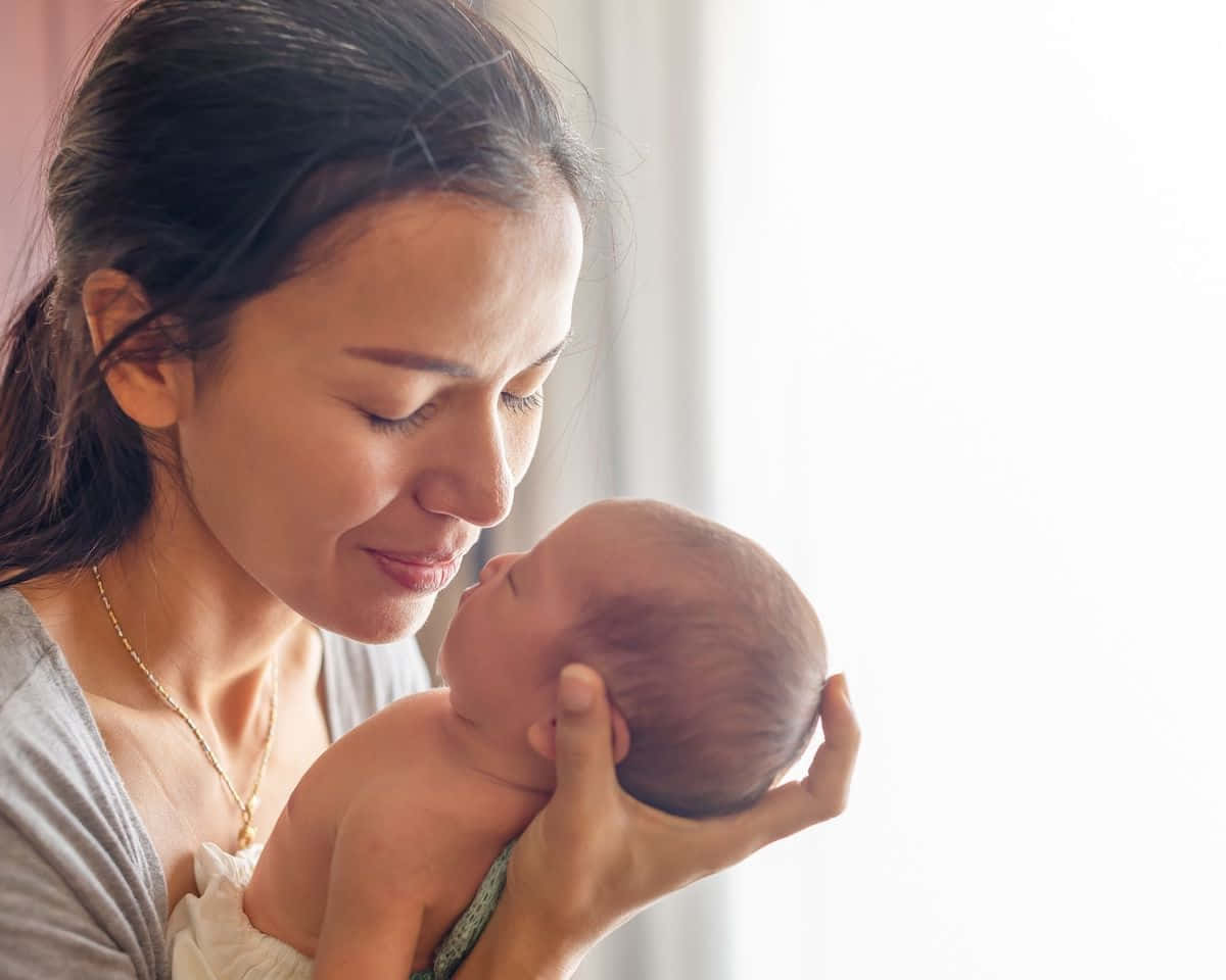 Newborn Baby 桌布 壁紙 — 一個溫柔的時刻，母親在柔和自然光中輕輕將新生嬰兒抱近臉龐。