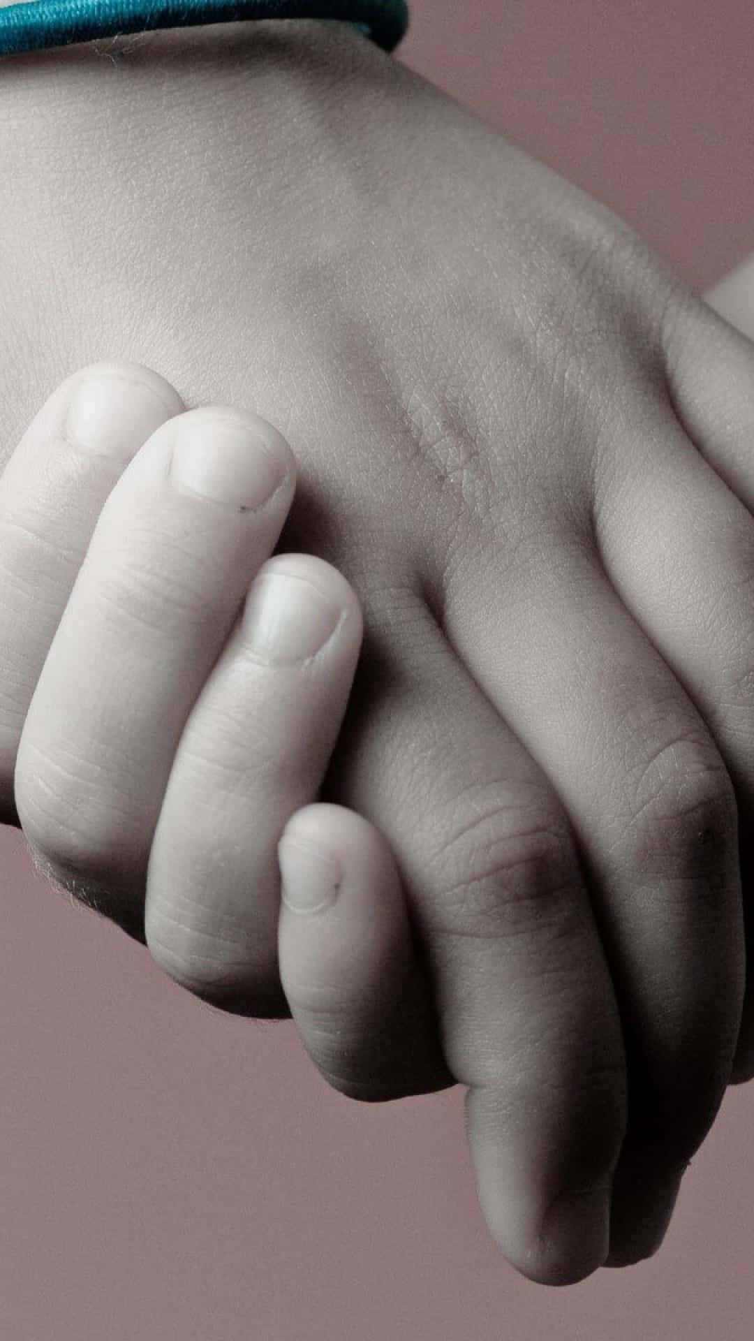 Holding Hands 桌布 壁紙 — 一個溫柔的特寫鏡頭，小孩的小手輕輕握住大人的大手，背景是柔和的粉紅色。
