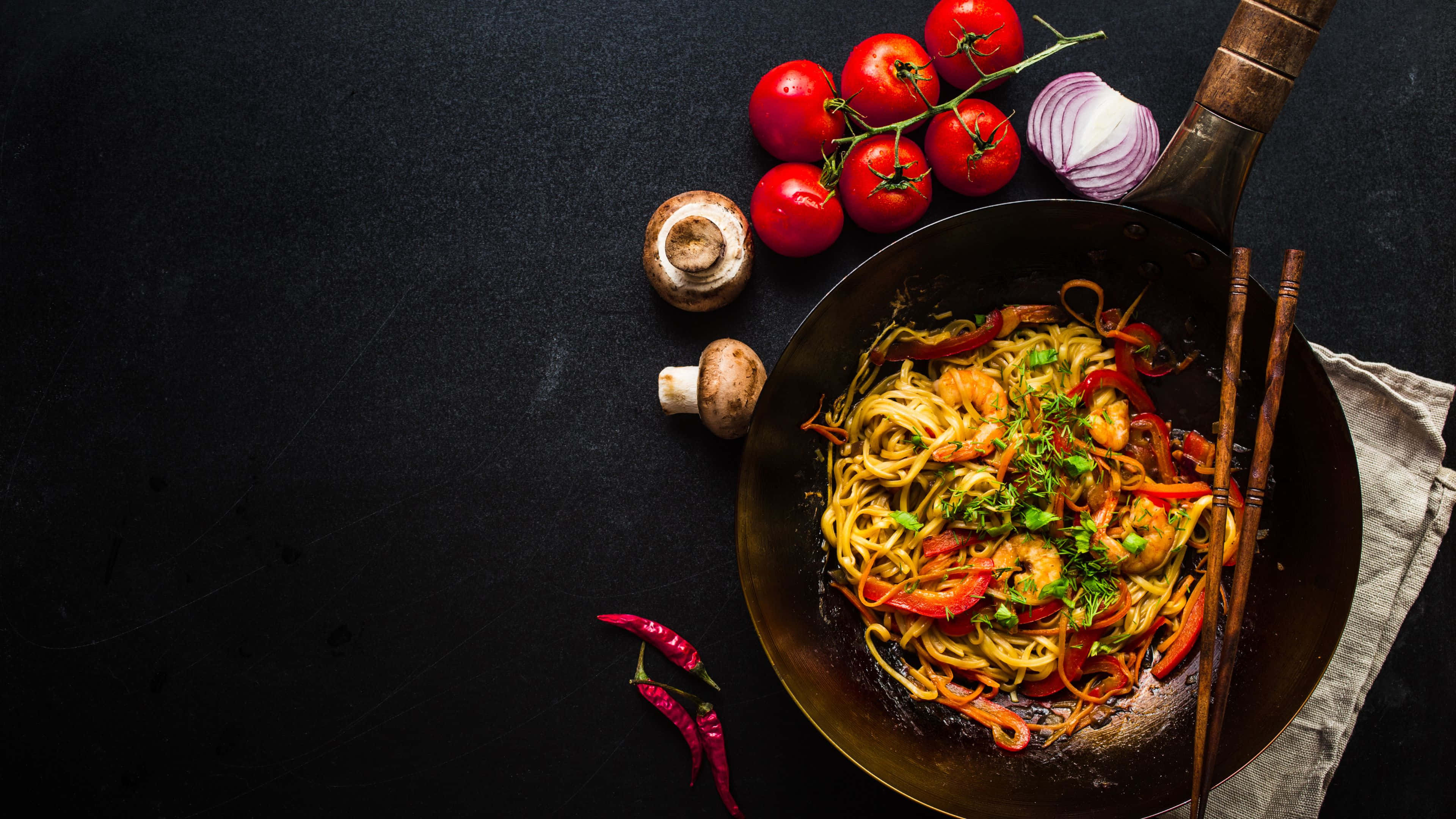 Noodles 桌布 壁紙 — 一個滋滋作響的炒鍋，裡面有色彩繽紛的炒麵，搭配蝦子、甜椒和香草，周圍是新鮮番茄、蘑菇和辣椒，放在深色表面上。