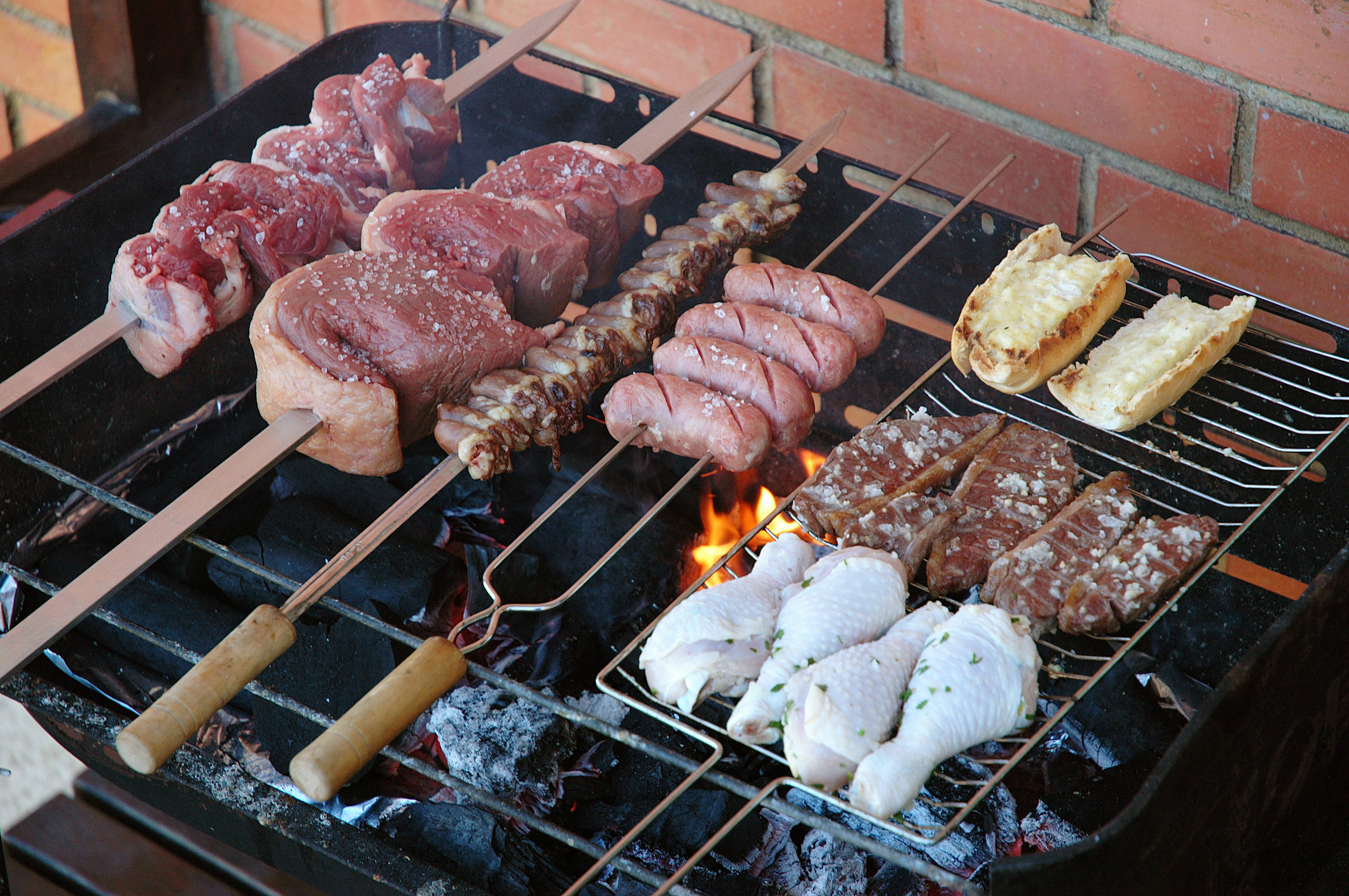 meat 桌布 壁紙 — 一個滋滋作響的燒烤架，上面放滿了串燒肉、香腸、雞肉和麵包，在熾熱的炭火上，背景是磚牆。
