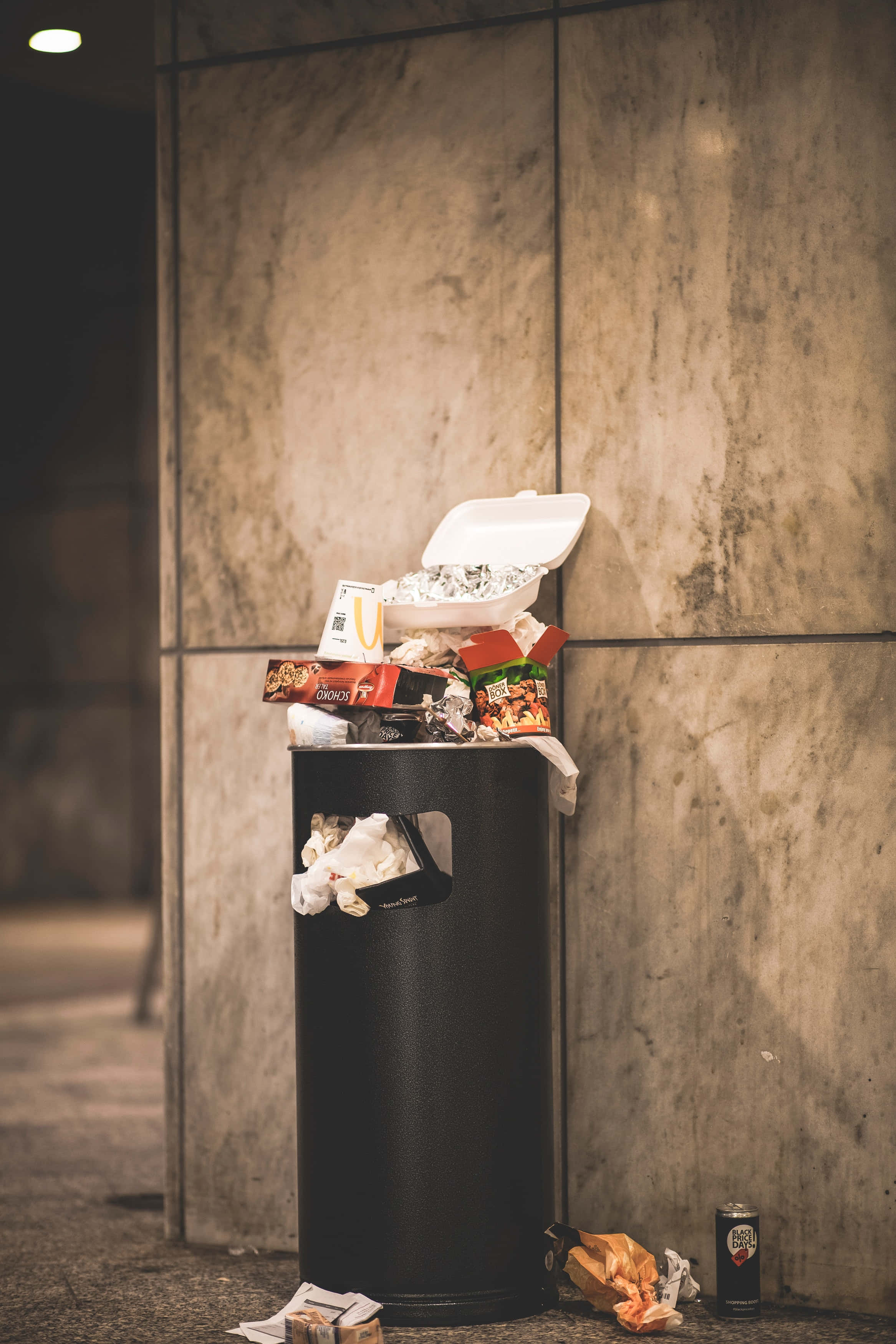 waste 桌布 壁紙 — 一個滿溢的黑色垃圾桶，裝滿速食包裝和保麗龍容器，靠在有紋理的石牆上，在夜晚。