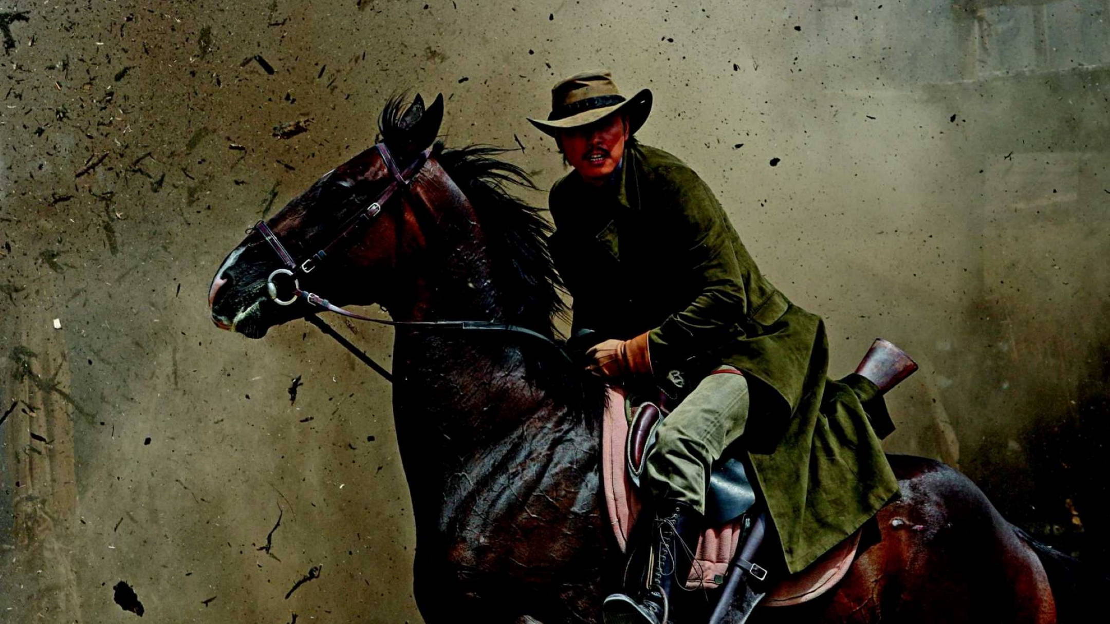 Cowboy 桌布 壁紙 — 一個滿臉是血的粗獷牛仔騎著一匹黑馬穿過塵土飛揚、碎片飛舞的惡劣環境。