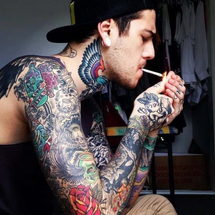 Tattoo Boy 桌布 壁紙 — 一個滿身刺青、戴黑色帽子的男人，在昏暗的房間裡，專注地點起一支煙。