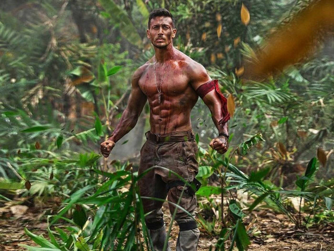 Indian 桌布 壁紙 — 一個滿身鮮血和泥濘的肌肉男在茂密的叢林中挑釁地站立，樹葉在他周圍旋轉。