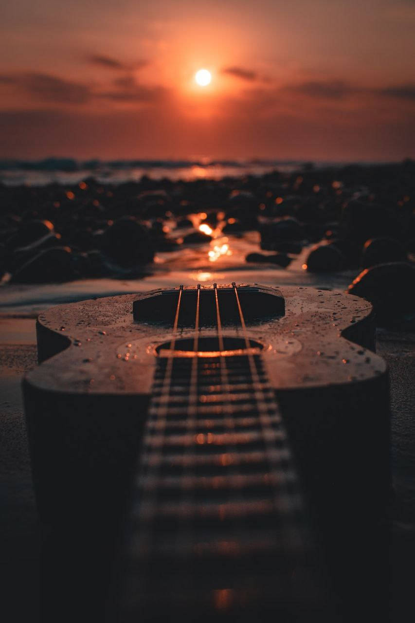 Beautiful Phone 桌布 壁紙 — 一個潮濕的烏克麗麗放在岩石海灘上，夕陽的溫暖橙色光線反射在水面和天空中。