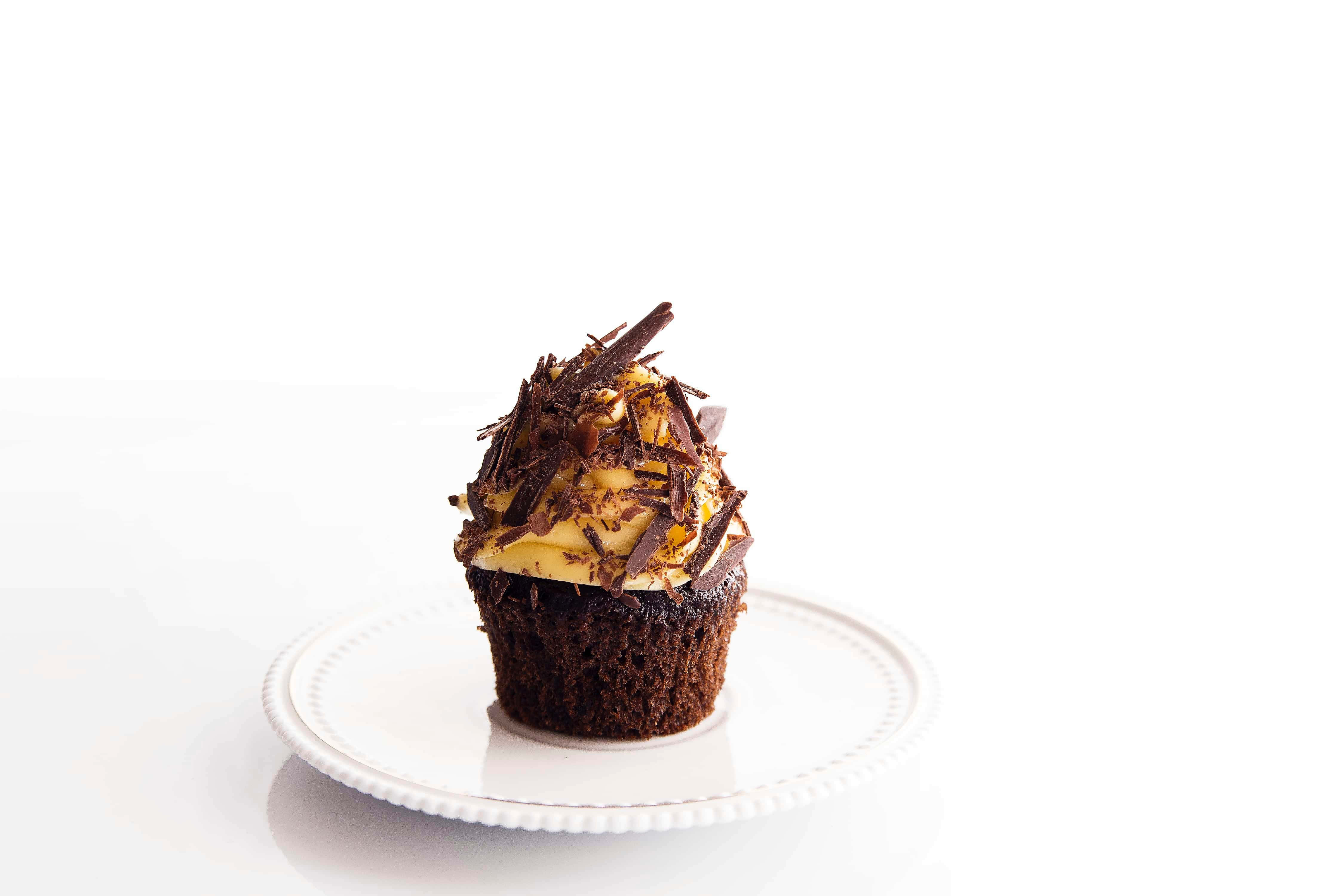 Cupcake 桌布 壁紙 — 一個濃郁的巧克力杯子蛋糕，上面有奶油黃色糖霜和黑巧克力碎，優雅地放在白色盤子上，背景是潔淨的白色。