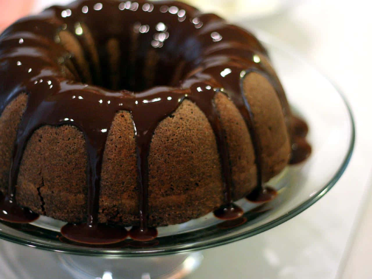 Chocolate Cake 桌布 壁紙 — 一個濃郁的巧克力環形蛋糕閃耀著光澤的巧克力糖霜，沿著其凹槽邊滴落到玻璃蛋糕架上。