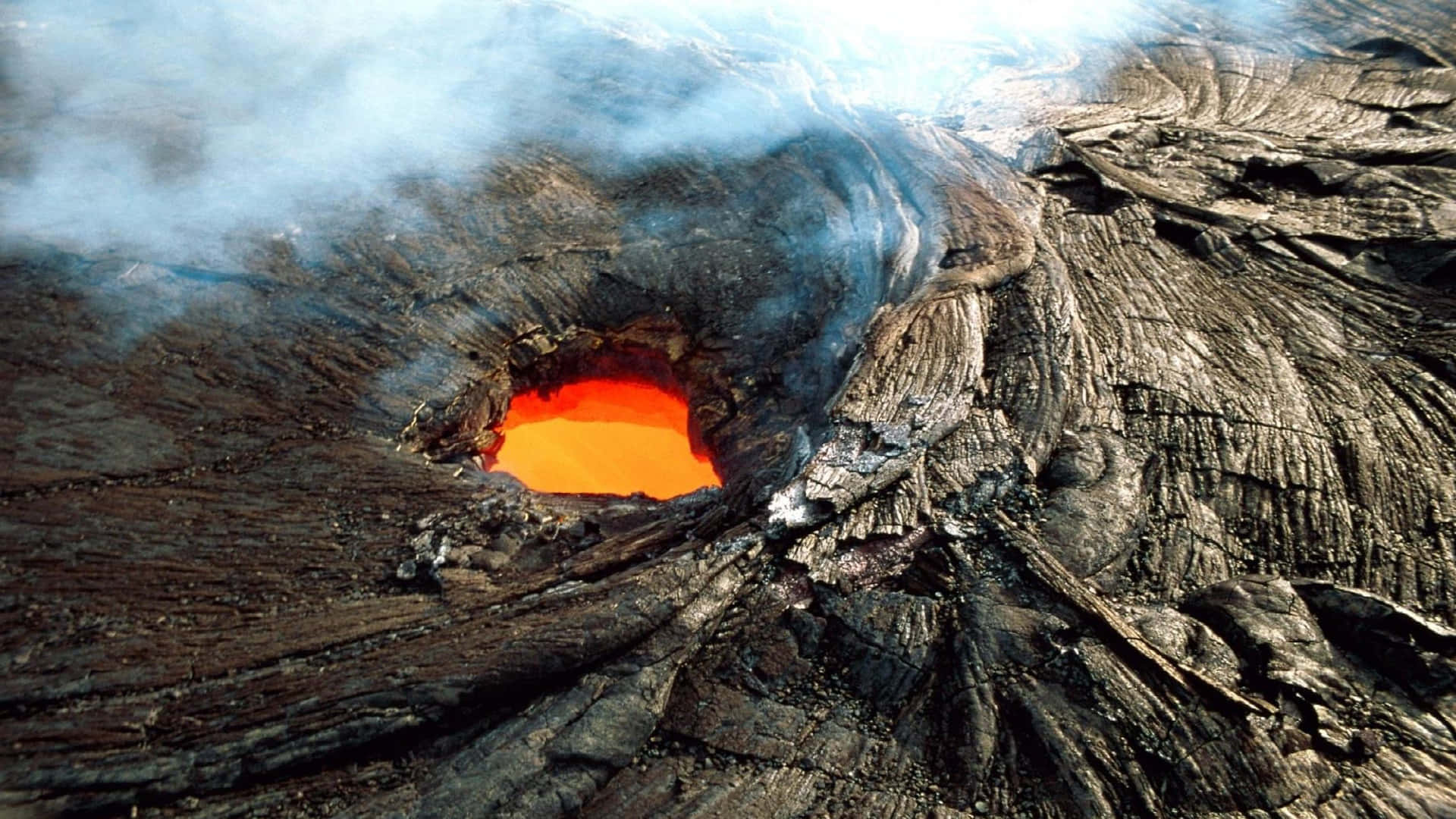 Volcano 桌布 壁紙 — 一個火山口噴發出熾熱的橙色熔岩，周圍環繞著崎嶇的黑色岩石和滾滾白煙。