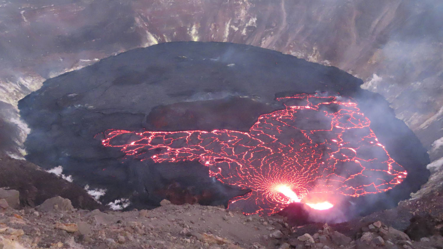 Volcano 桌布 壁紙 — 一個火山口發出鮮紅色的熔岩光芒，流過黑暗崎嶇的地形，天空朦朧。