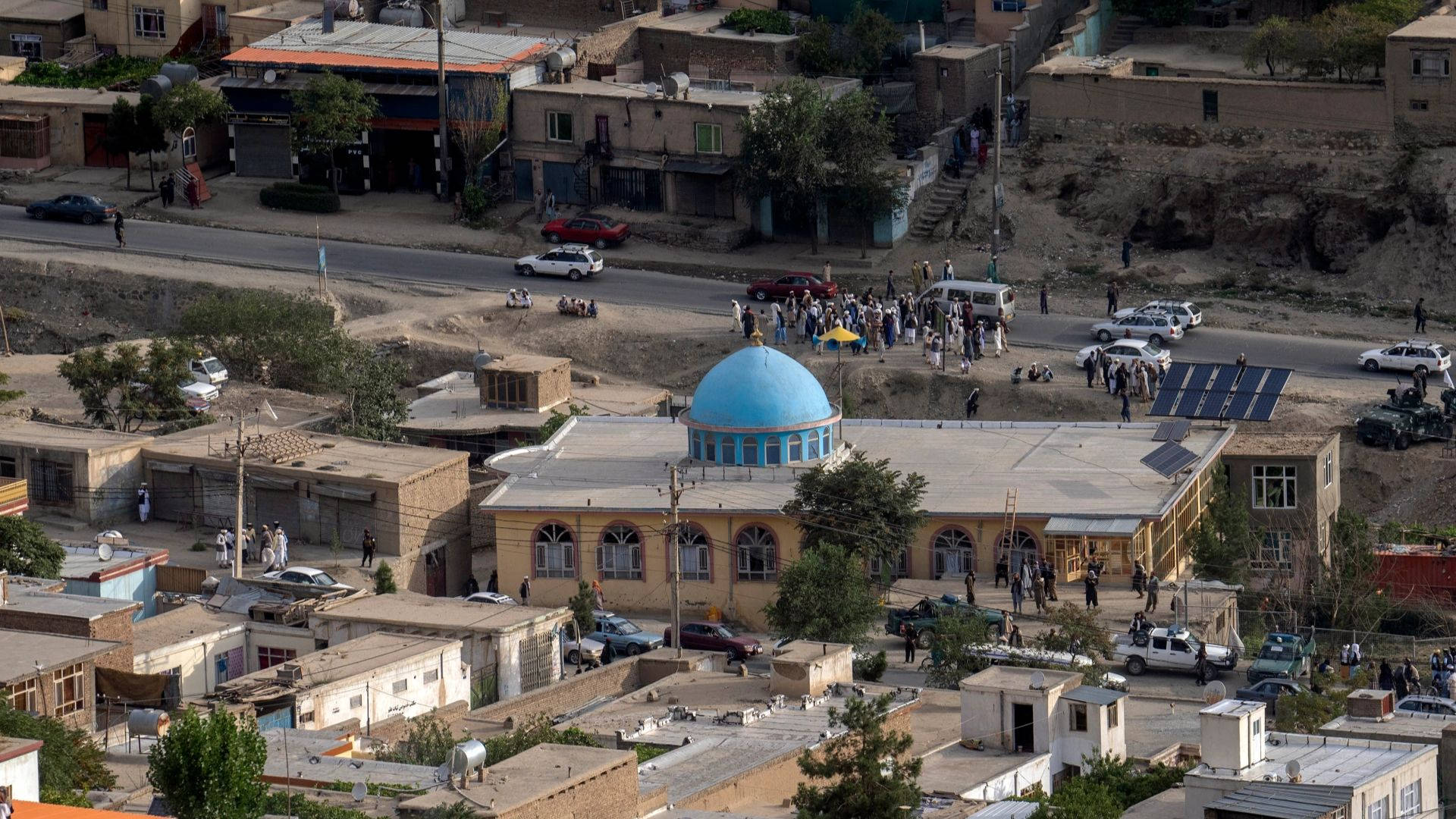 Eritrea 桌布 壁紙 — 一個灰塵飛揚的小鎮的鳥瞰圖，有一座藍色圓頂清真寺，周圍是低矮建築，靠近一條有車輛的道路旁聚集著人群。