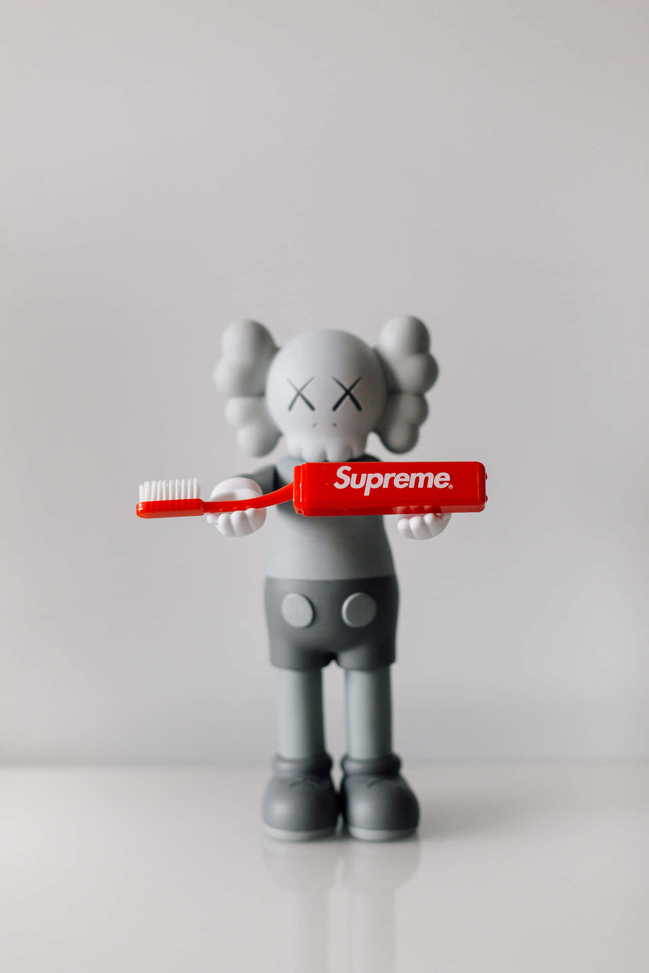 Kaws X Supreme 桌布 壁紙 — 一個灰色KAWS公仔在簡約白色背景前拿著紅色Supreme牙刷。