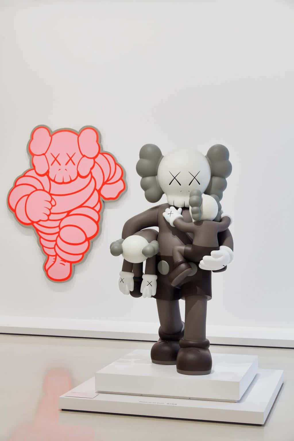 Kaws Figures 桌布 壁紙 — 一個灰色KAWS雕塑手持一個較小的公仔，站在白色底座上，旁邊是畫廊牆上一個粉紅色輪廓的KAWS藝術品。