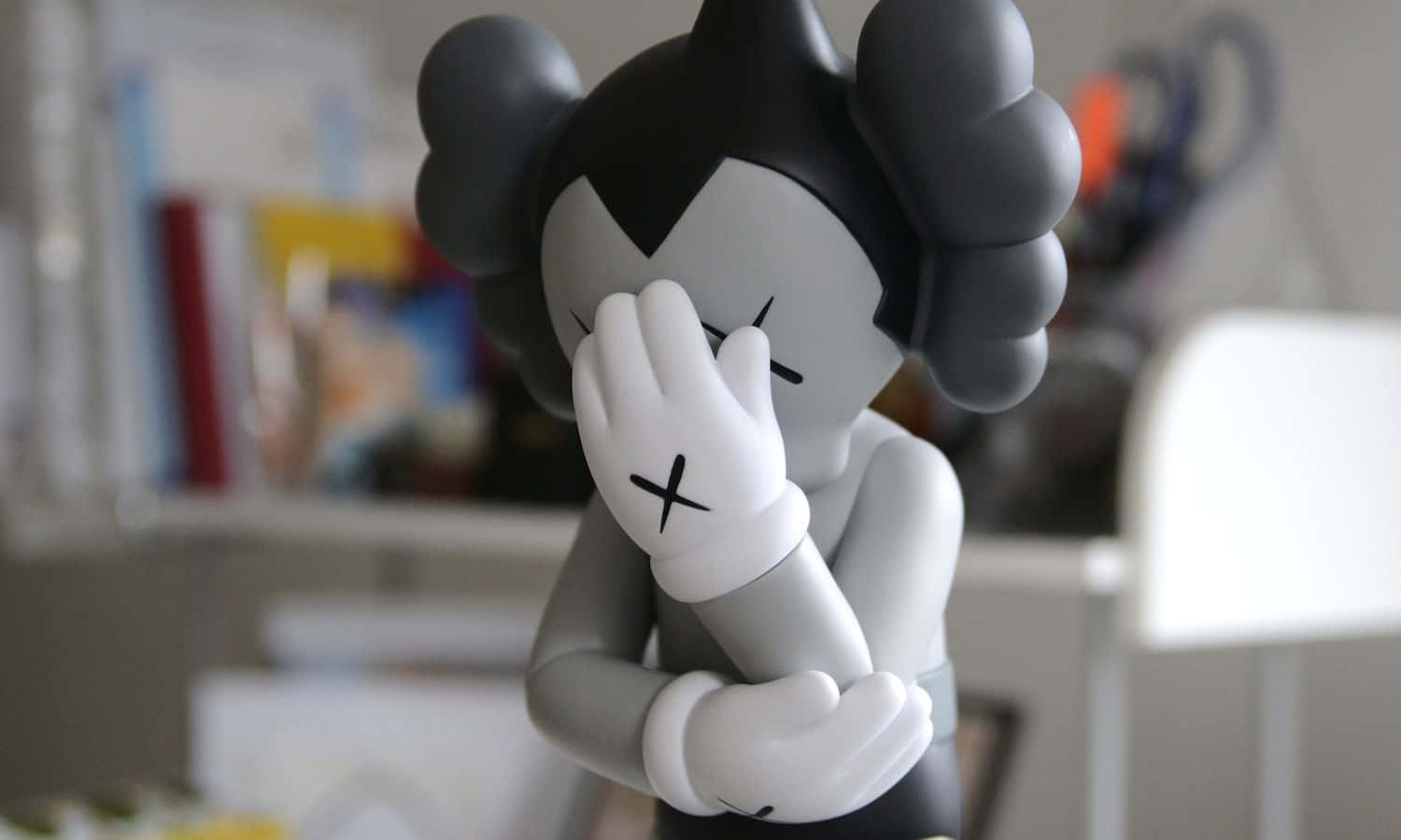 Kaws Astro Boy 桌布 壁紙 — 一個灰色和白色的KAWS公仔，黑色頭髮遮住臉部，坐在模糊的室內環境中。