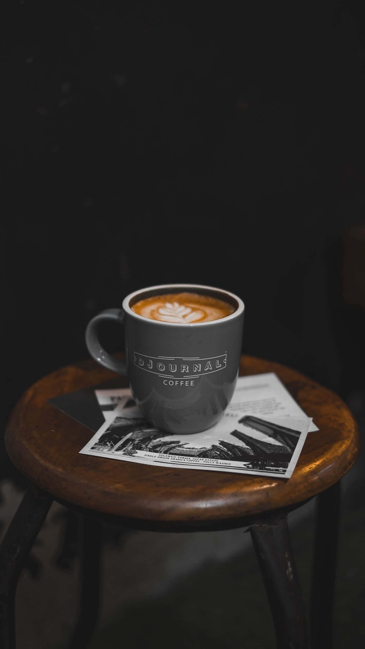 Coffee Aesthetic 桌布 壁紙 — 一個灰色咖啡杯帶有拉花藝術，放在木凳上，置於報紙之上，處於昏暗的環境中。