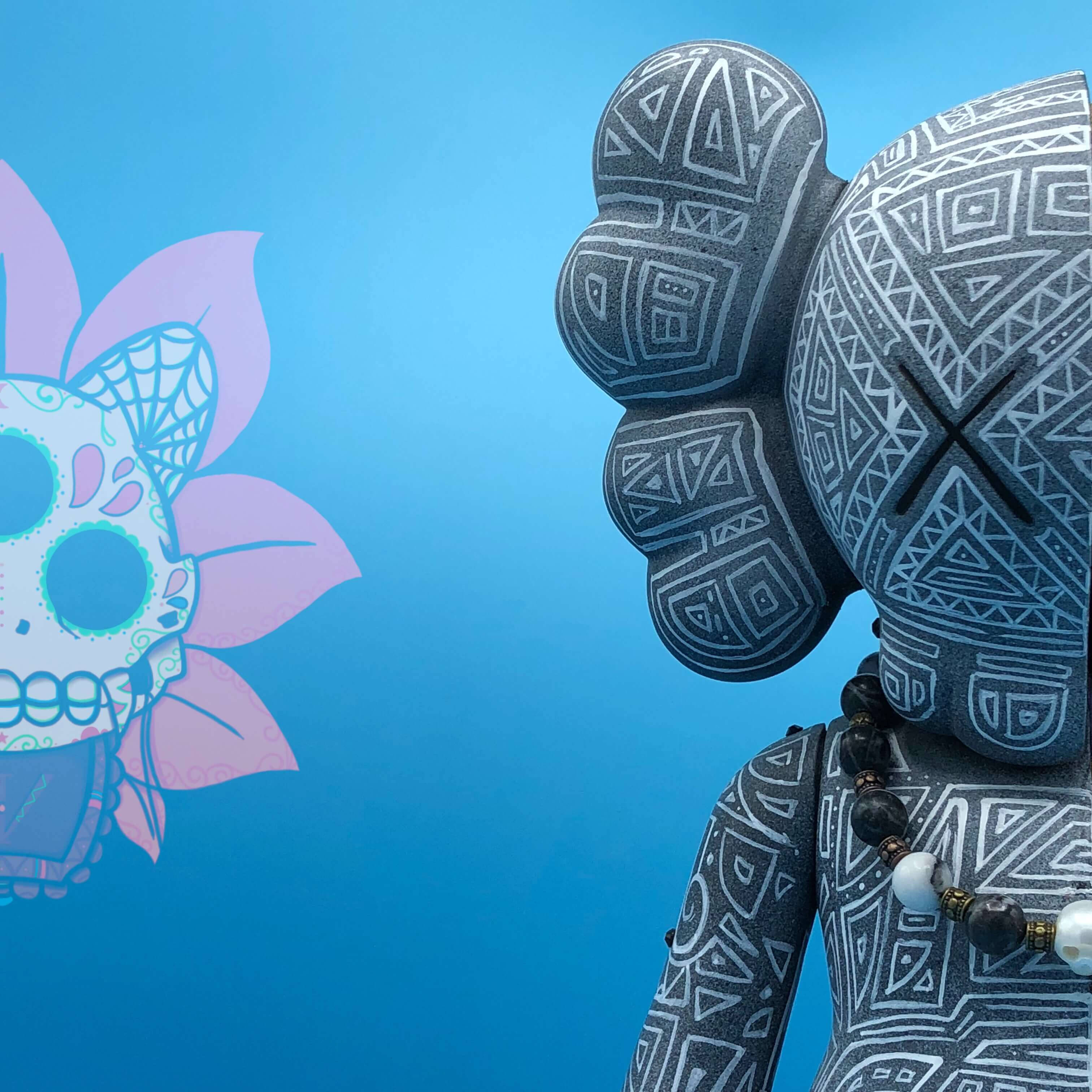 KAWS 桌布 壁紙 — 一個灰色圖案KAWS公仔，帶有黑色X眼睛，在純藍色背景上凝視著一個色彩繽紛的糖骷髏花旁邊。