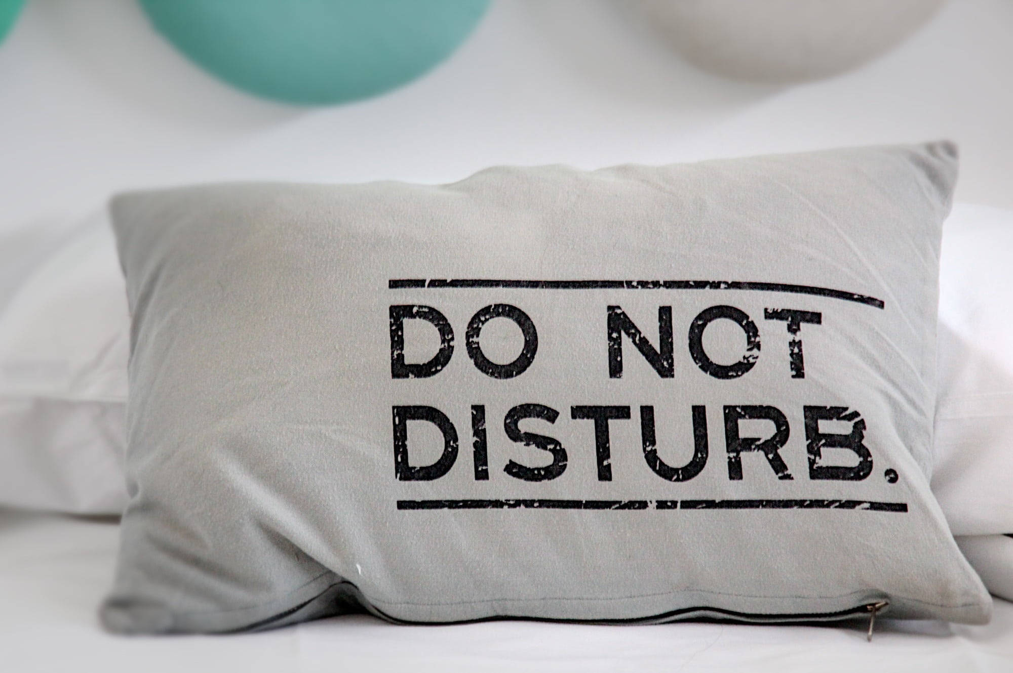 搞笑 桌布 壁紙 — 一個灰色枕頭，上面有粗體黑色「DO NOT DISTURB」字樣，放在一張鋪有柔軟白色床單的床上，背景有青綠色氣球。
