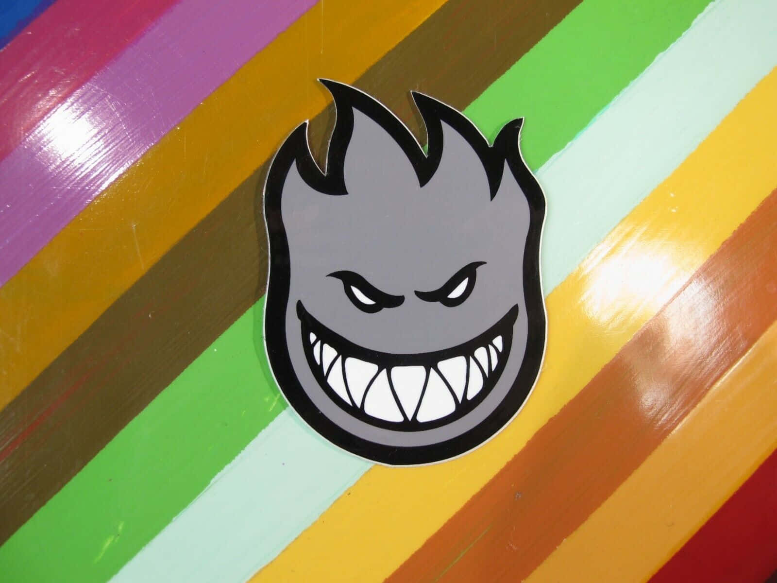 Spitfire Skate 桌布 壁紙 — 一個灰色火焰頭貼紙，帶著咧嘴笑的表情，貼在光滑的彩虹條紋表面上。