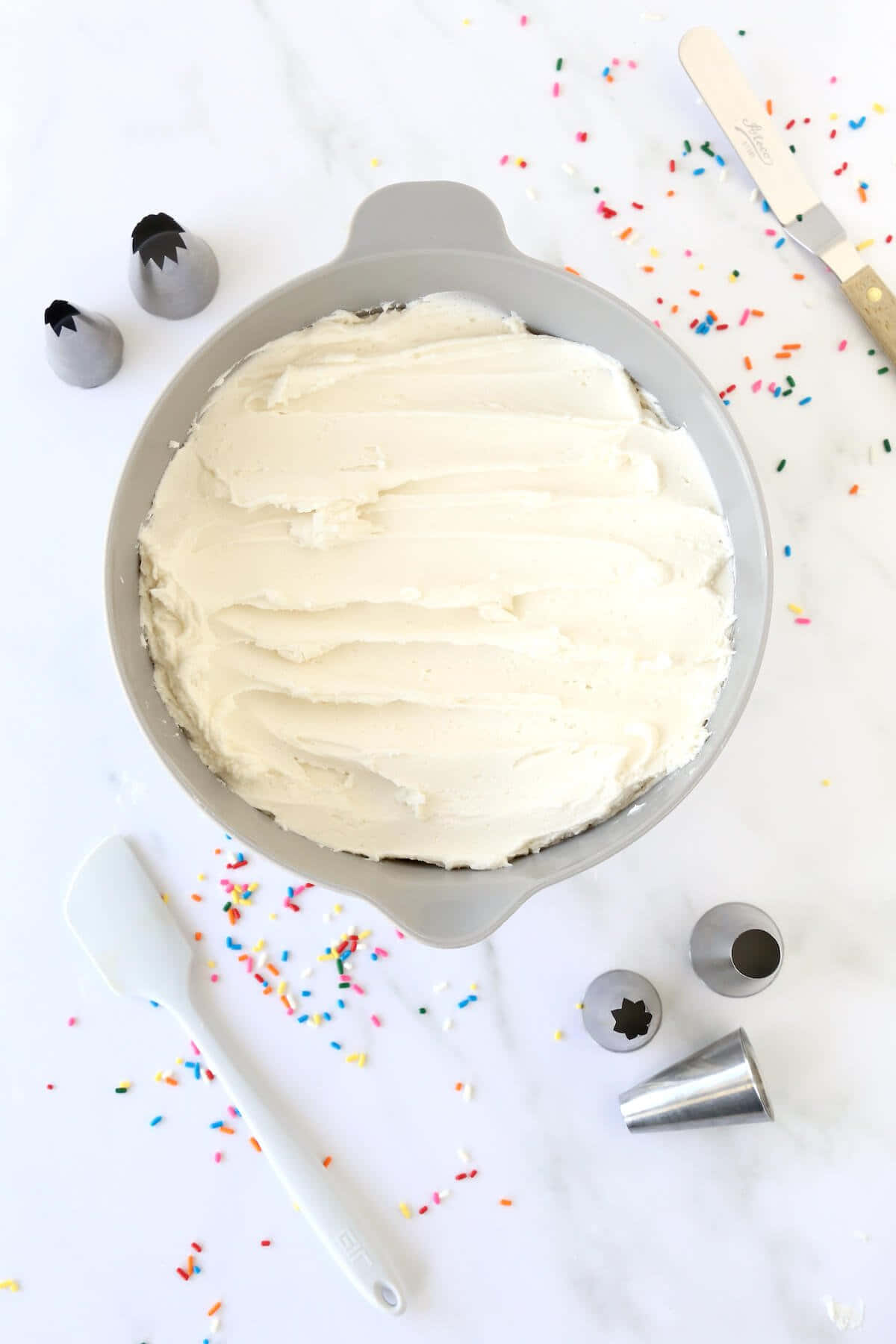 Buttercream 桌布 壁紙 — 一個灰色烤盤中的糖霜蛋糕，周圍是彩色糖粒和裱花嘴，置於大理石檯面上。