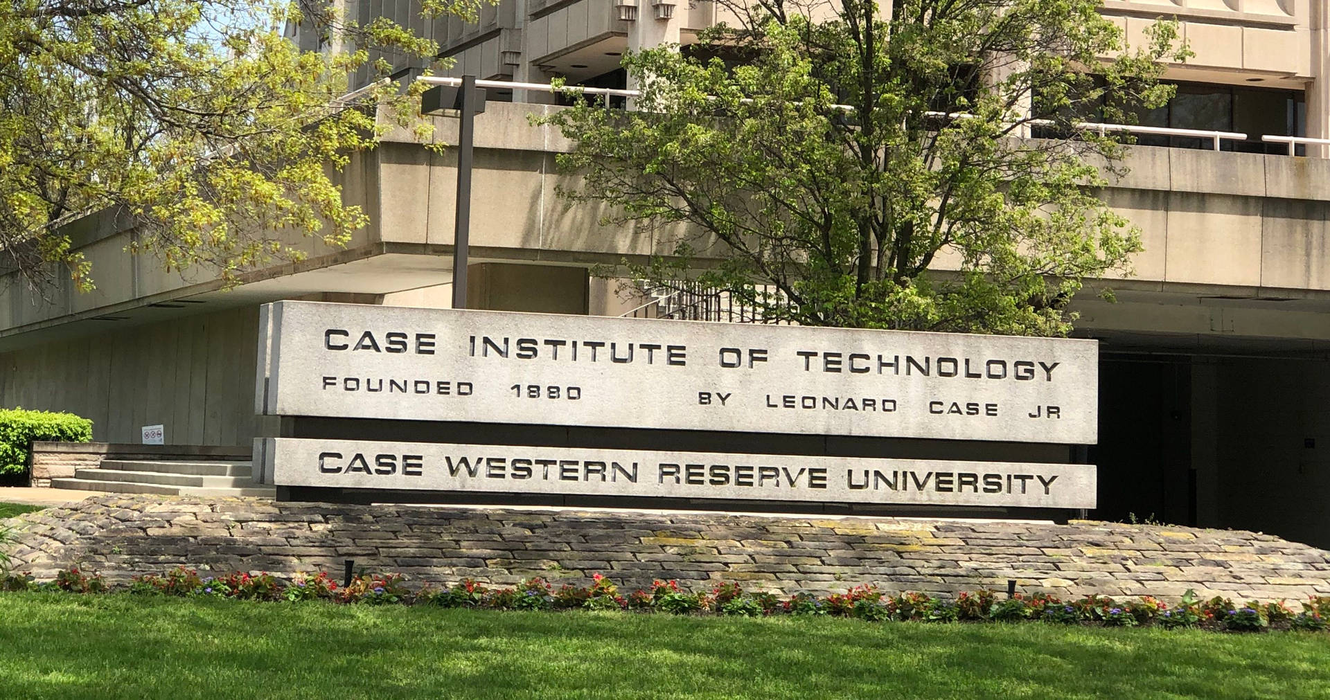 壁紙 — 一個為凱斯理工學院和凱斯西儲大學而設的石製標誌，矗立在一座現代建築前，周圍有綠樹和草地，在明亮的日光下。