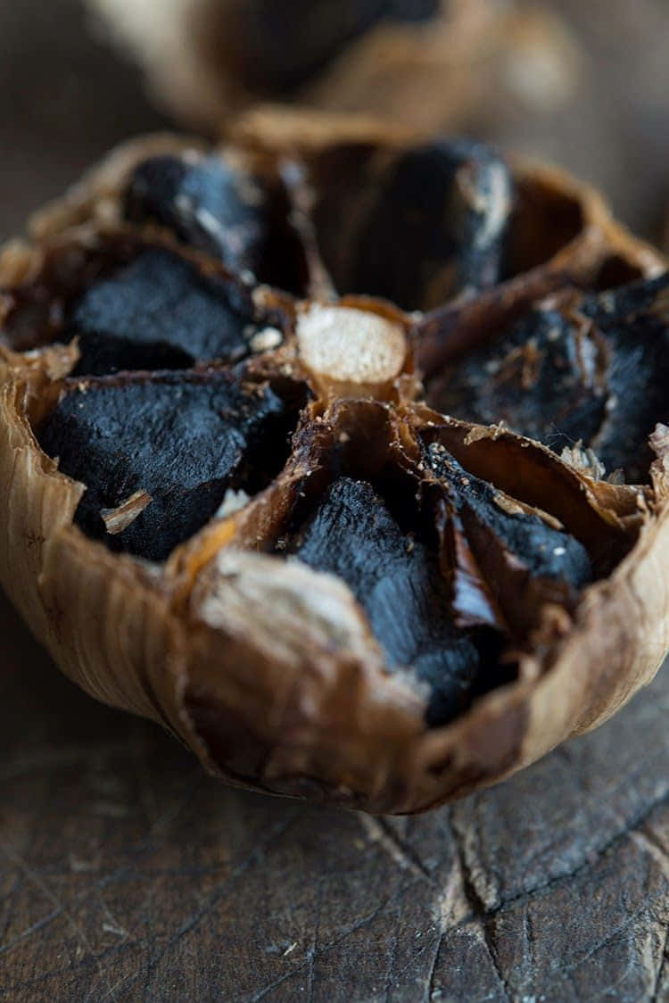 Black Garlic 桌布 壁紙 — 一個烤大蒜頭被掰開的特寫，在質樸的木質表面上露出燒焦的黑色蒜瓣。