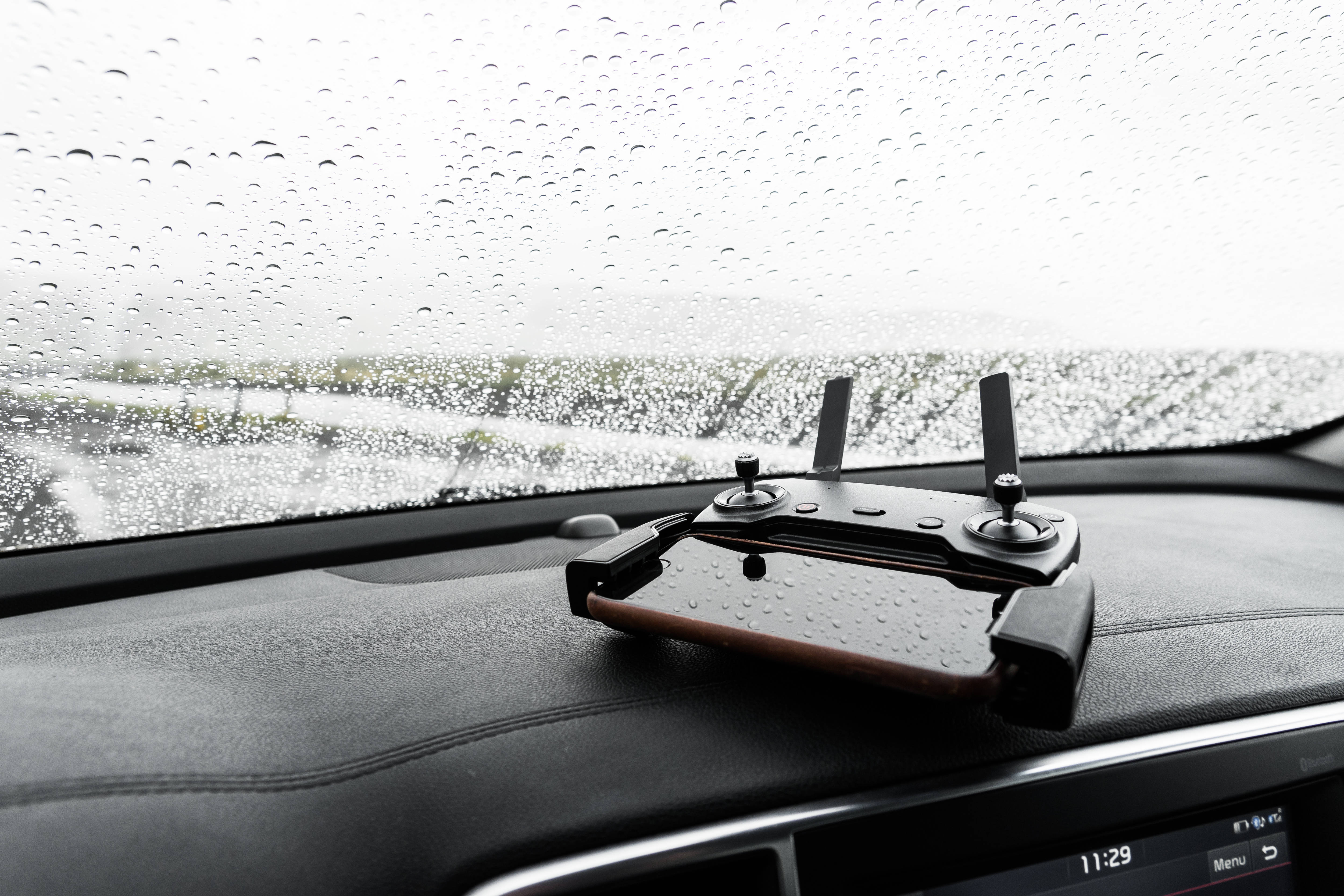 Raining 桌布 壁紙 — 一個無人機控制器放在潮濕的汽車儀表板上，外面的擋風玻璃上有雨滴劃過。