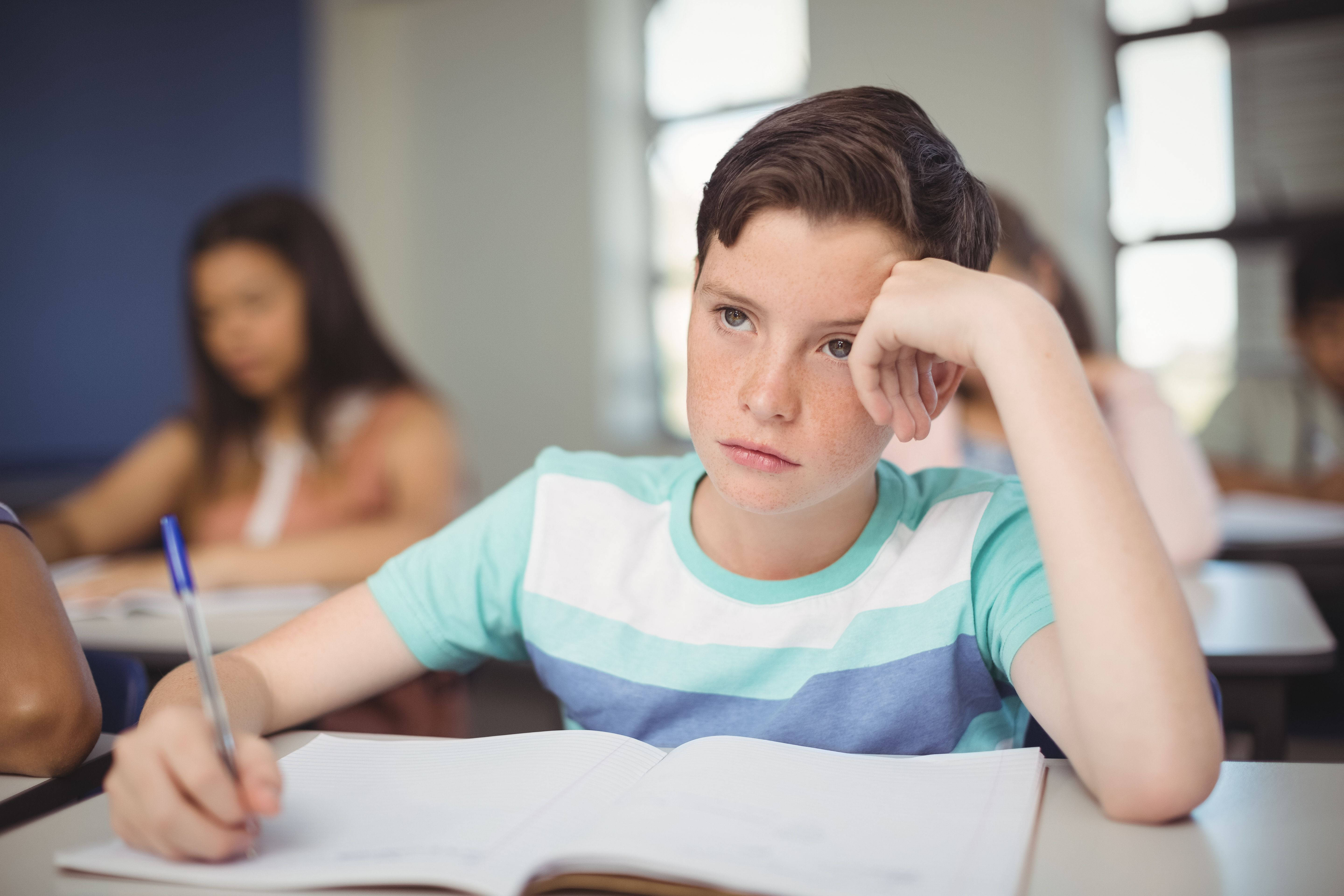 書桌 桌布 壁紙 — 一個無聊的青少年男孩將頭靠在手上，同時在教室裡握著一支筆放在打開的筆記本上。