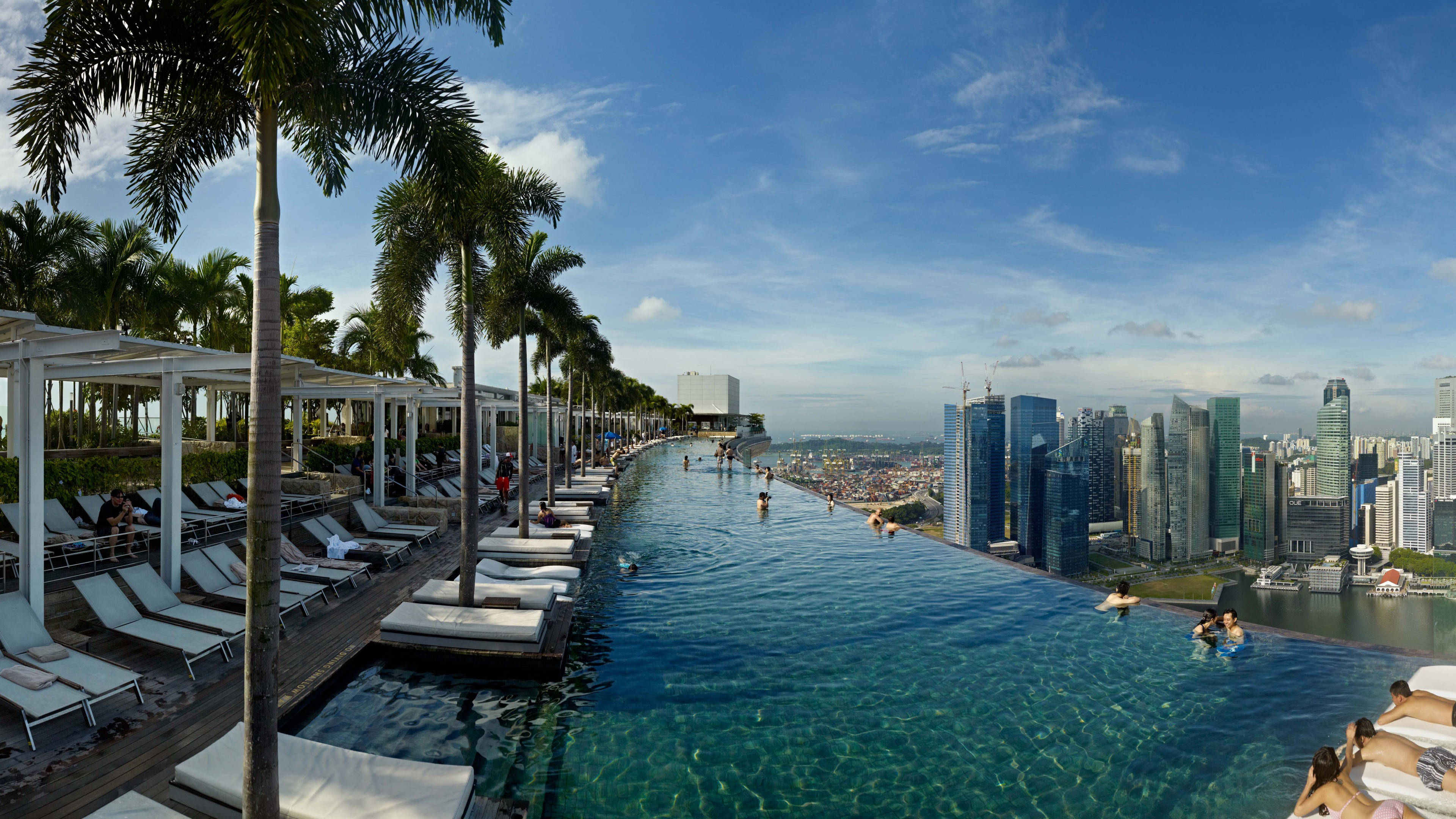 Singapore 桌布 壁紙 — 一個無邊際泳池俯瞰現代城市天際線，棕櫚樹和躺椅在明亮的藍天下。