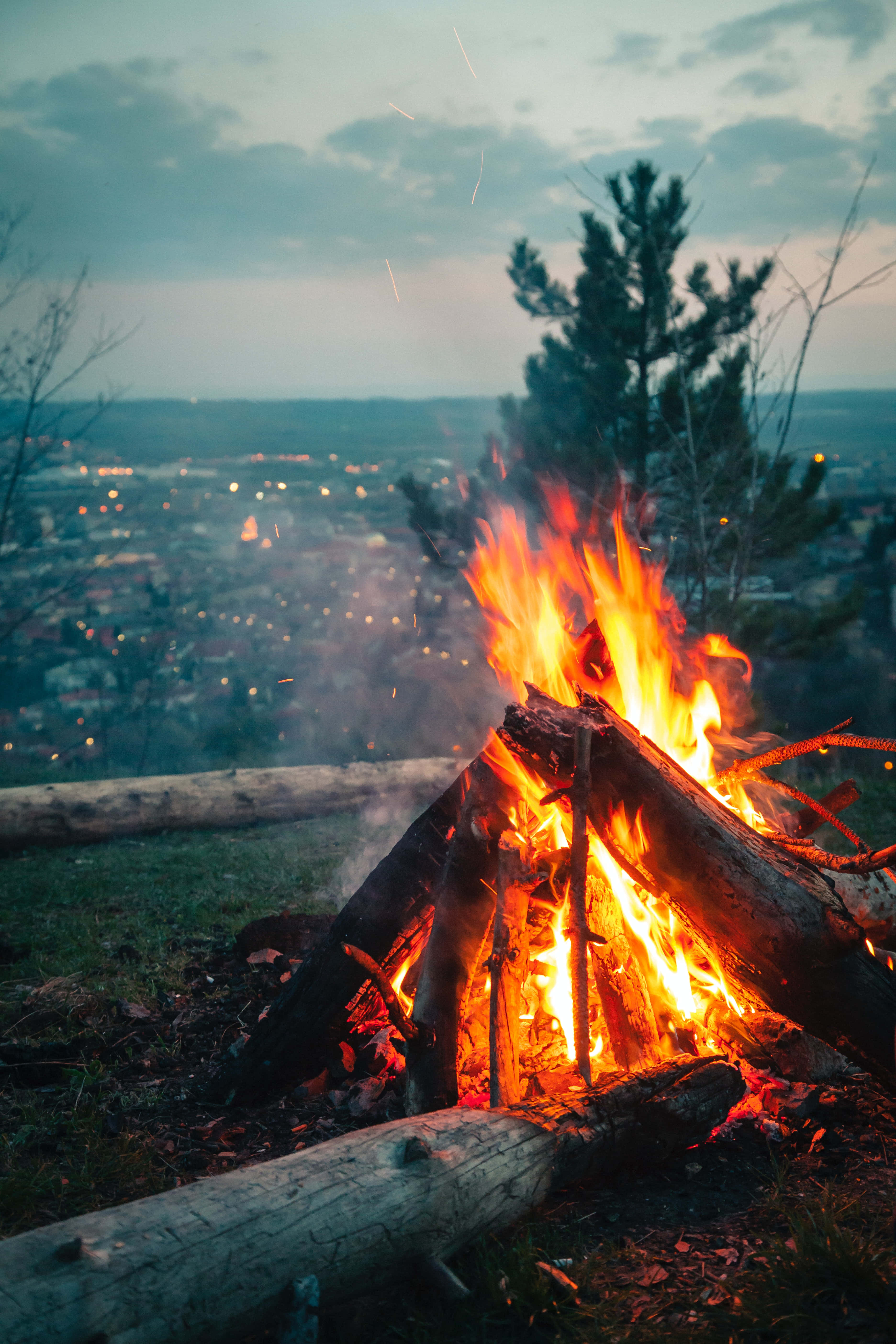 Bonfire 桌布 壁紙 — 一個熊熊燃燒的營火，帶著熾熱的餘燼和飛舞的火星，照亮了一片俯瞰遠處城市的草地山坡，此時正值黃昏。