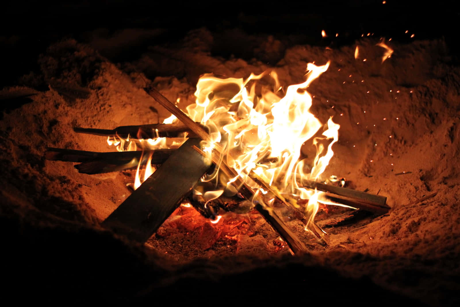 Beach Bonfire 桌布 壁紙 — 一個熊熊燃燒的營火在夜間的雪地中明亮地燃燒，將溫暖的橙色光芒投射到周圍的雪堆上。