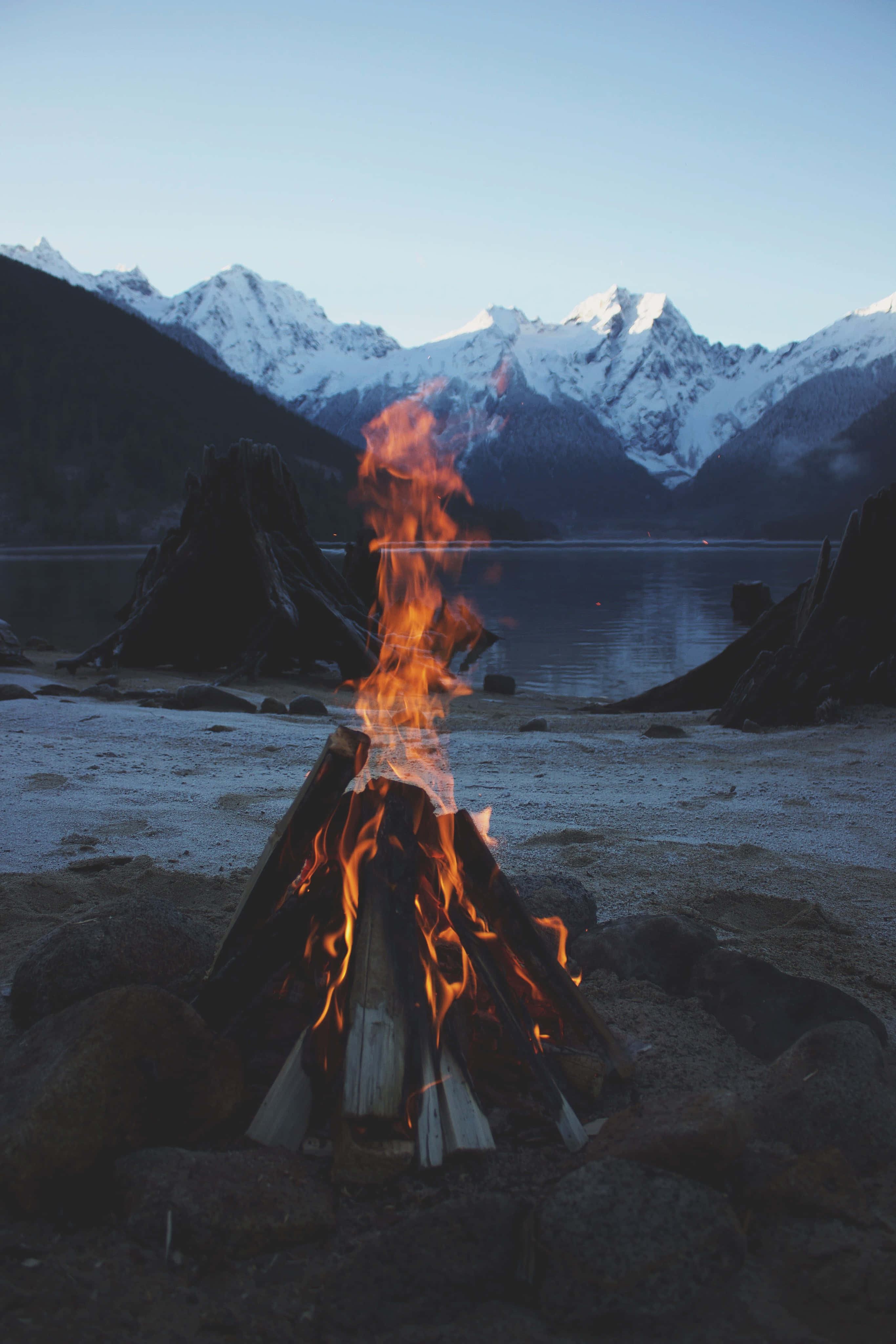 Bonfire 桌布 壁紙 — 一個熊熊燃燒的營火在白雪覆蓋的湖岸上明亮地燃燒，積雪覆蓋的山脈在暮色中升起。