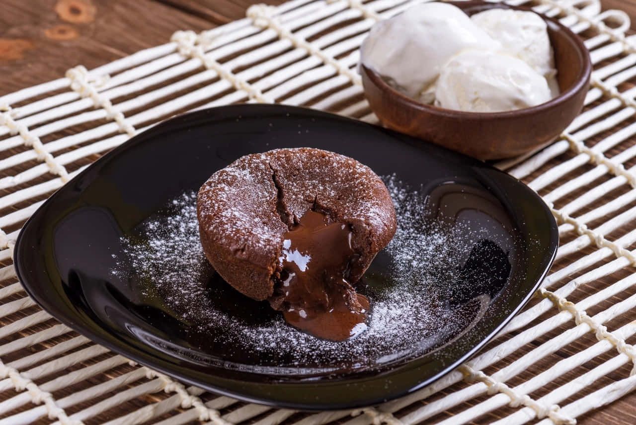 Winter Desserts 桌布 壁紙 — 一個熔岩巧克力蛋糕流出濃郁黏稠的巧克力醬到一個灑滿糖粉的黑色盤子上，旁邊放著一碗香草冰淇淋。