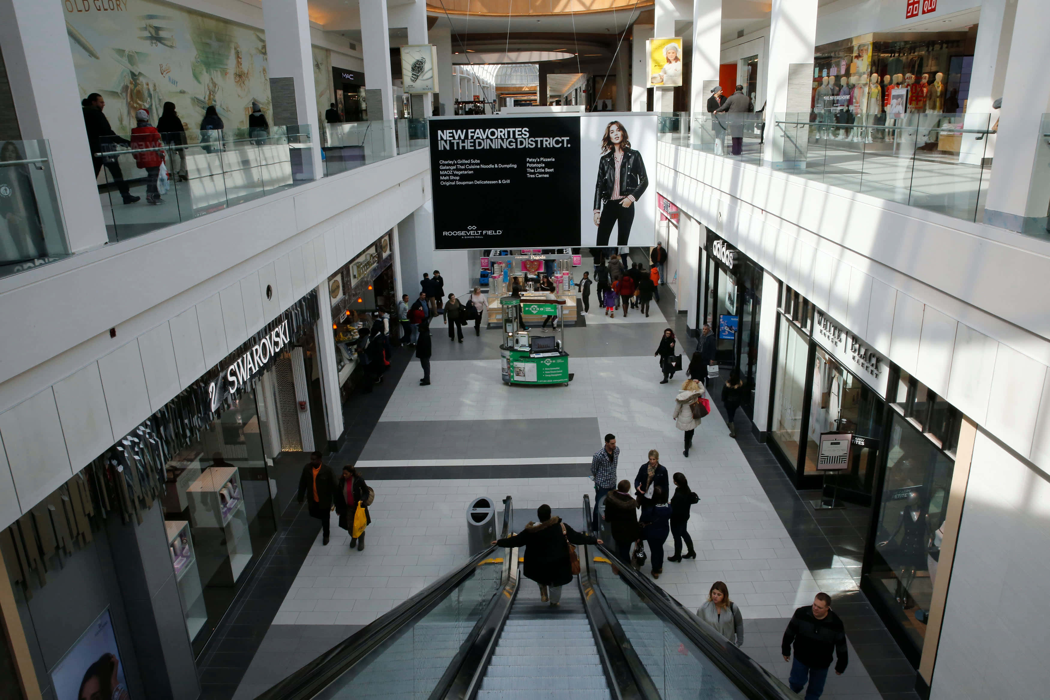 Mall 桌布 壁紙 — 一個熙熙攘攘的多層購物中心，購物者在明亮的自然光下行走並使用自動扶梯。