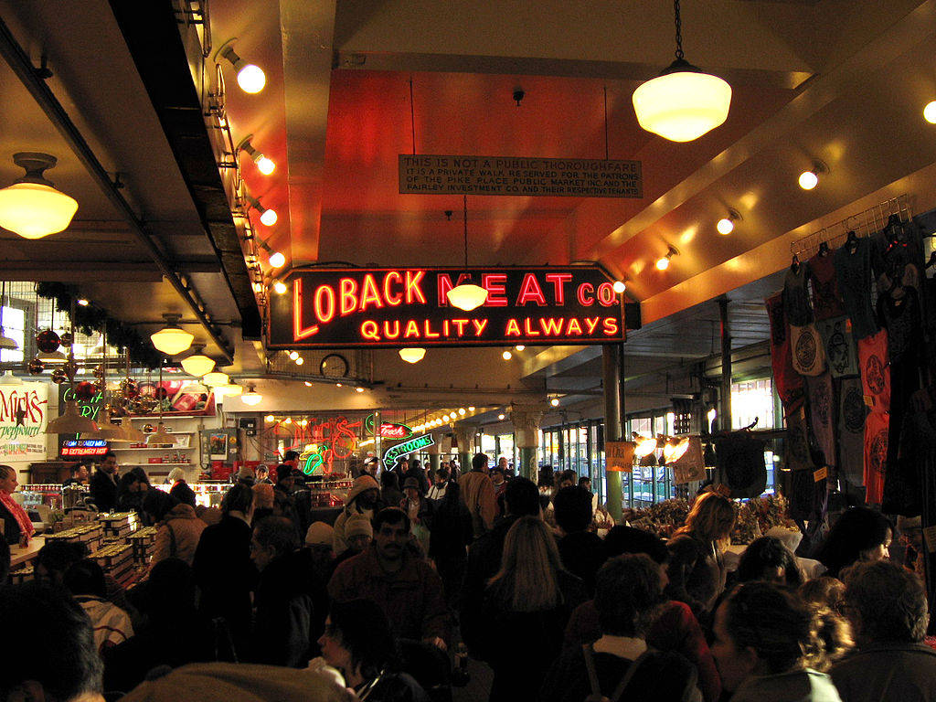 Pike Place Market 桌布 壁紙 — 一個熙熙攘攘的室內市場，在溫暖的吊燈下，一個發光的紅色霓虹燈招牌「Loback Meat Co」懸掛在購物者人群上方。