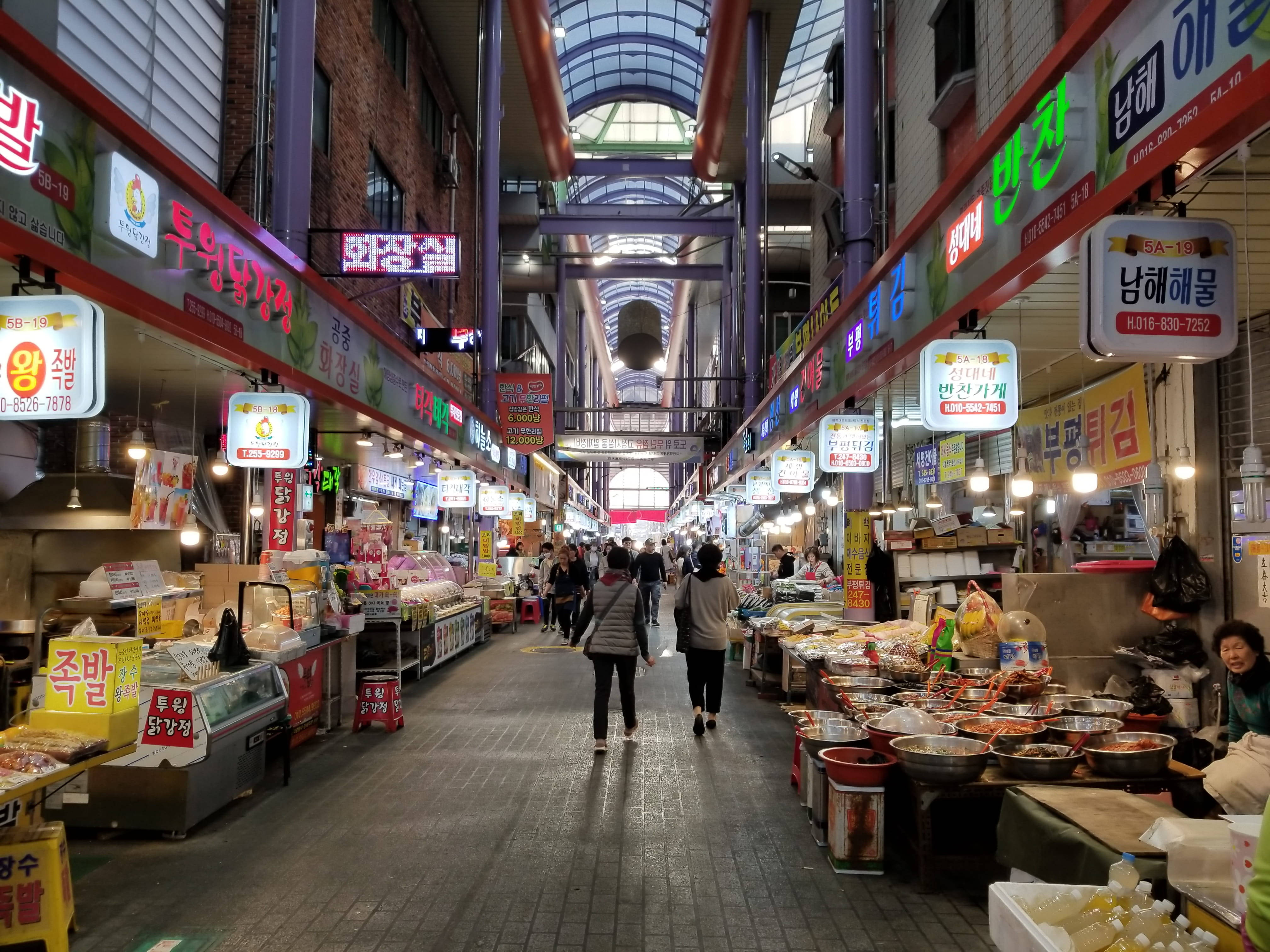 Busan 桌布 壁紙 — 一個熙熙攘攘的室內韓國市場，頭頂上有鮮豔的霓虹燈招牌，購物者在擺滿色彩繽紛食物的攤位之間穿梭。