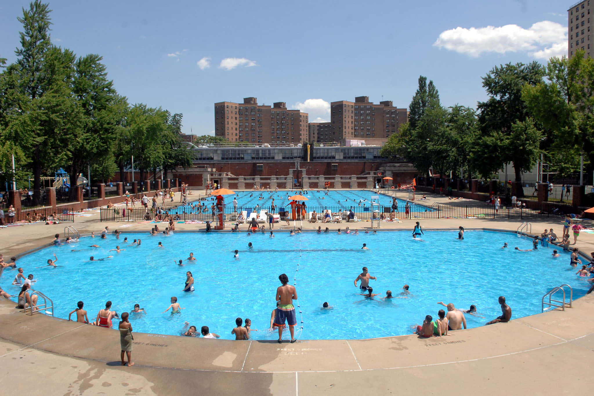 pool 桌布 壁紙 — 一個熙熙攘攘的戶外公眾游泳池，擠滿了游泳者，背景是明亮的藍天、散落的雲朵和公寓大樓。