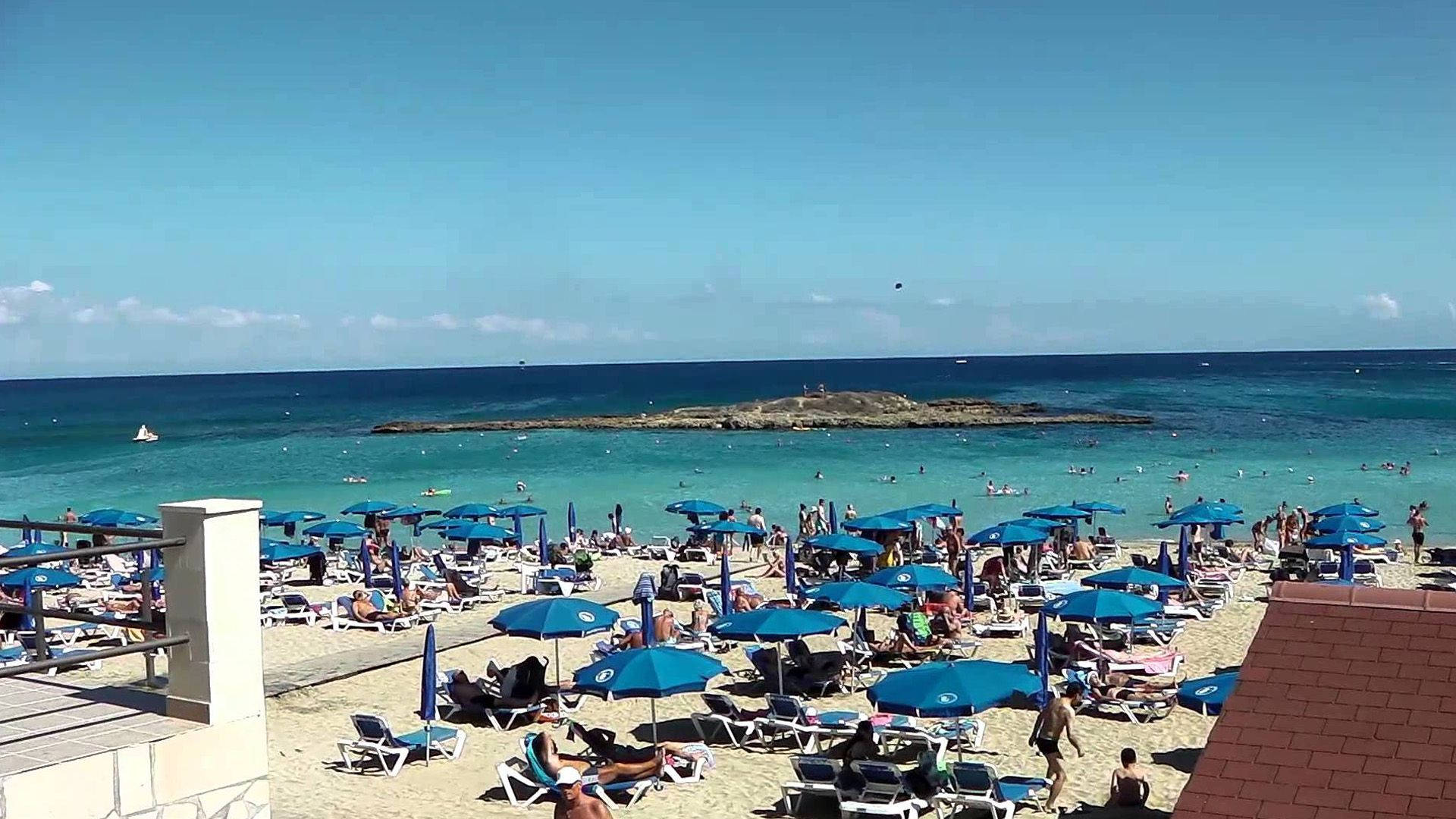 Cyprus 桌布 壁紙 — 一個熙熙攘攘的海灘，擠滿了在亮藍色遮陽傘下享受日光浴的人們，背景是碧綠的海水和晴朗的天空。