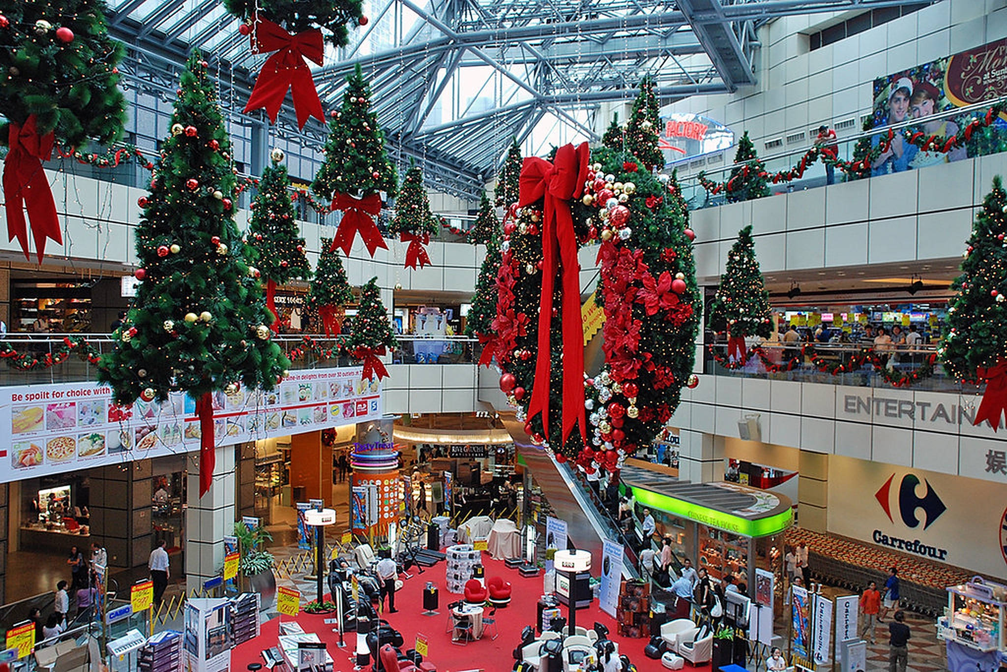Mall 桌布 壁紙 — 一個熙熙攘攘的購物中心中庭，裝飾著高聳的聖誕樹和從玻璃天花板垂下的紅色蝴蝶結。