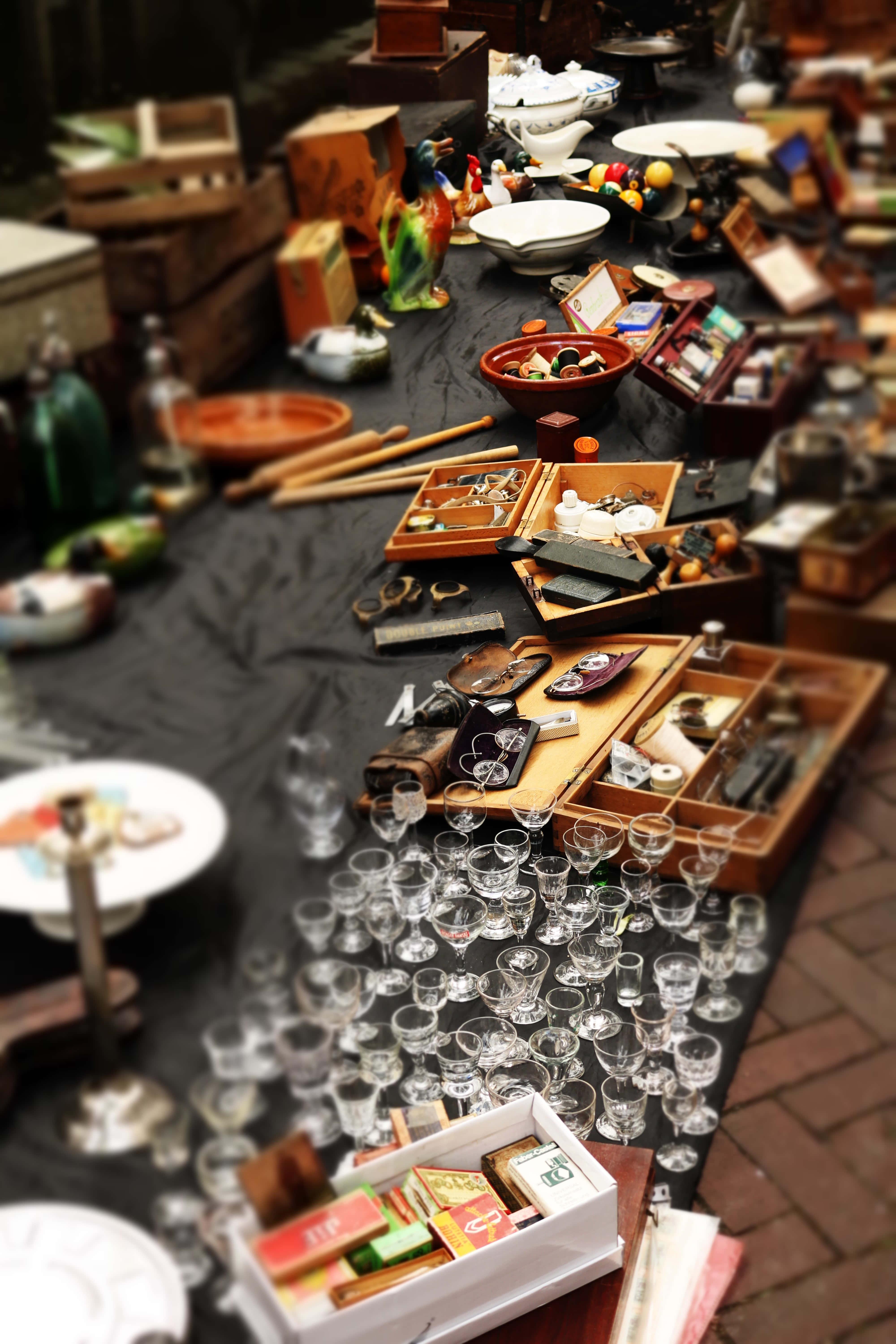 antique 桌布 壁紙 — 一個熙熙攘攘的跳蚤市場攤位，在柔和日光下堆滿了復古玻璃器皿、木箱和各式各樣的收藏品。