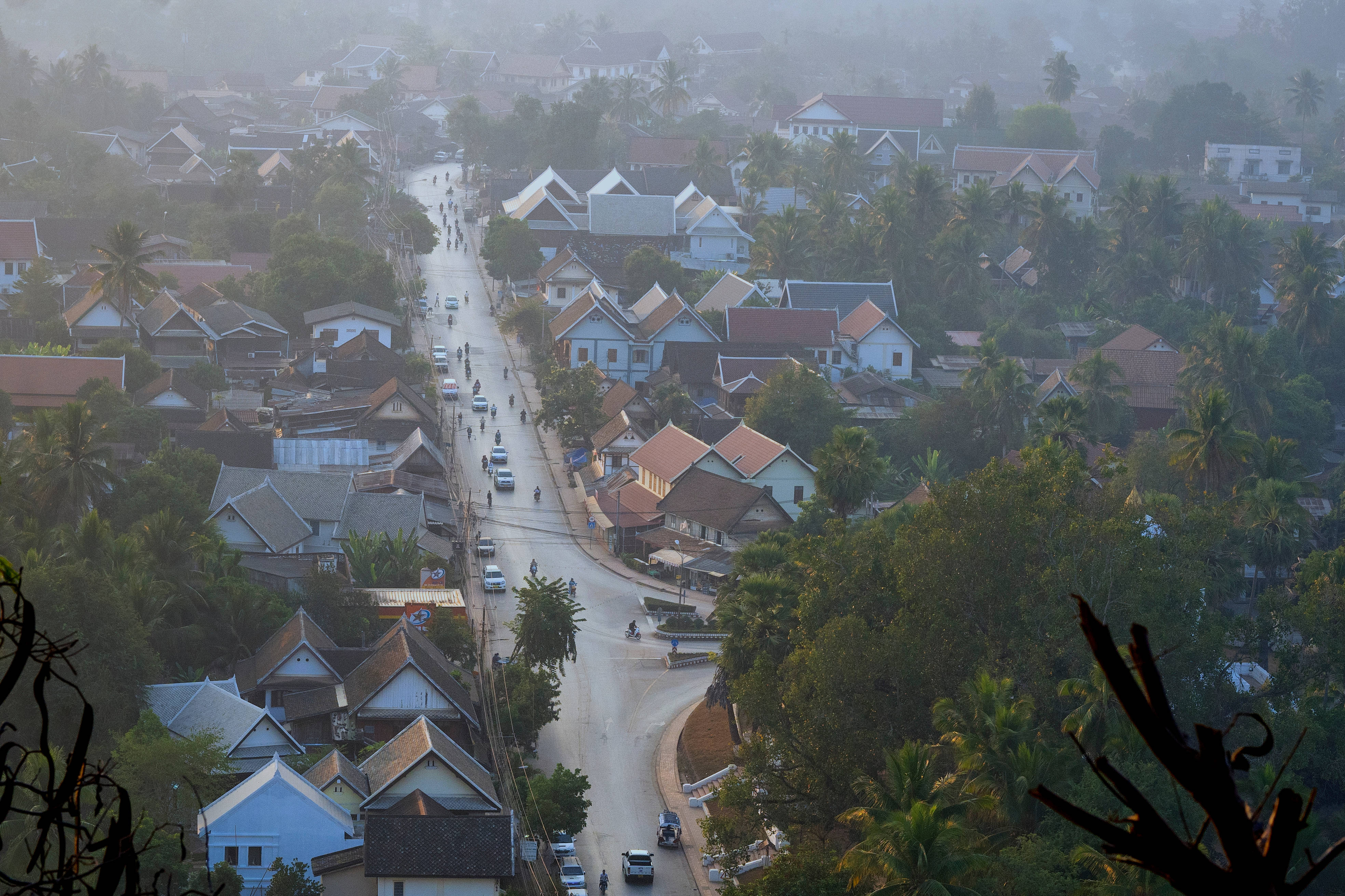 Laos 桌布 壁紙 — 一個熱帶村莊的霧氣黎明景色，蜿蜒的道路、色彩繽紛的房屋和棕櫚樹點綴在茂密的綠意之中。