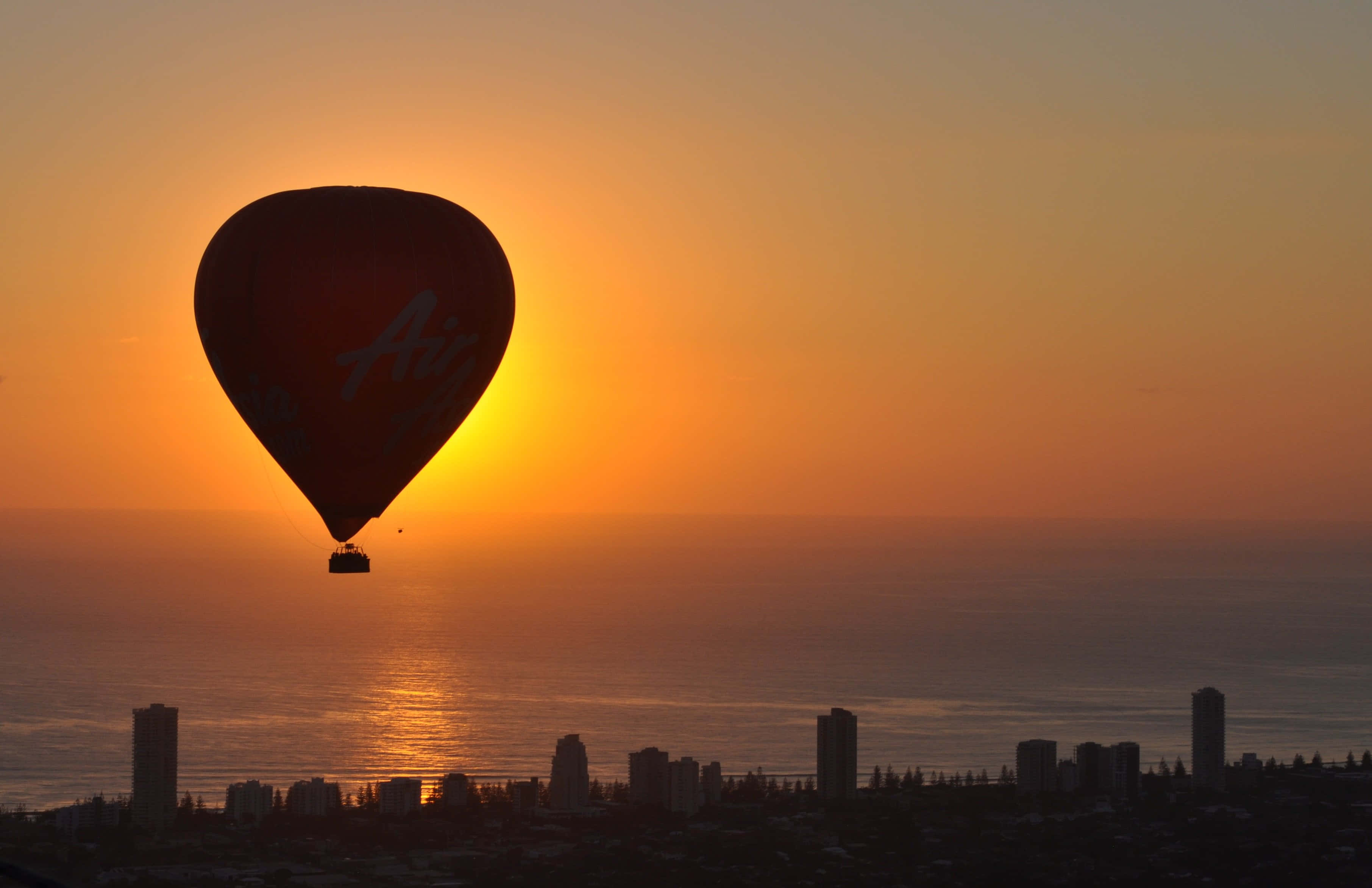 Hot Air Balloon 桌布 壁紙 — 一個熱氣球在火紅的日落映襯下形成剪影，飄浮在海洋上空，下方是城市天際線。