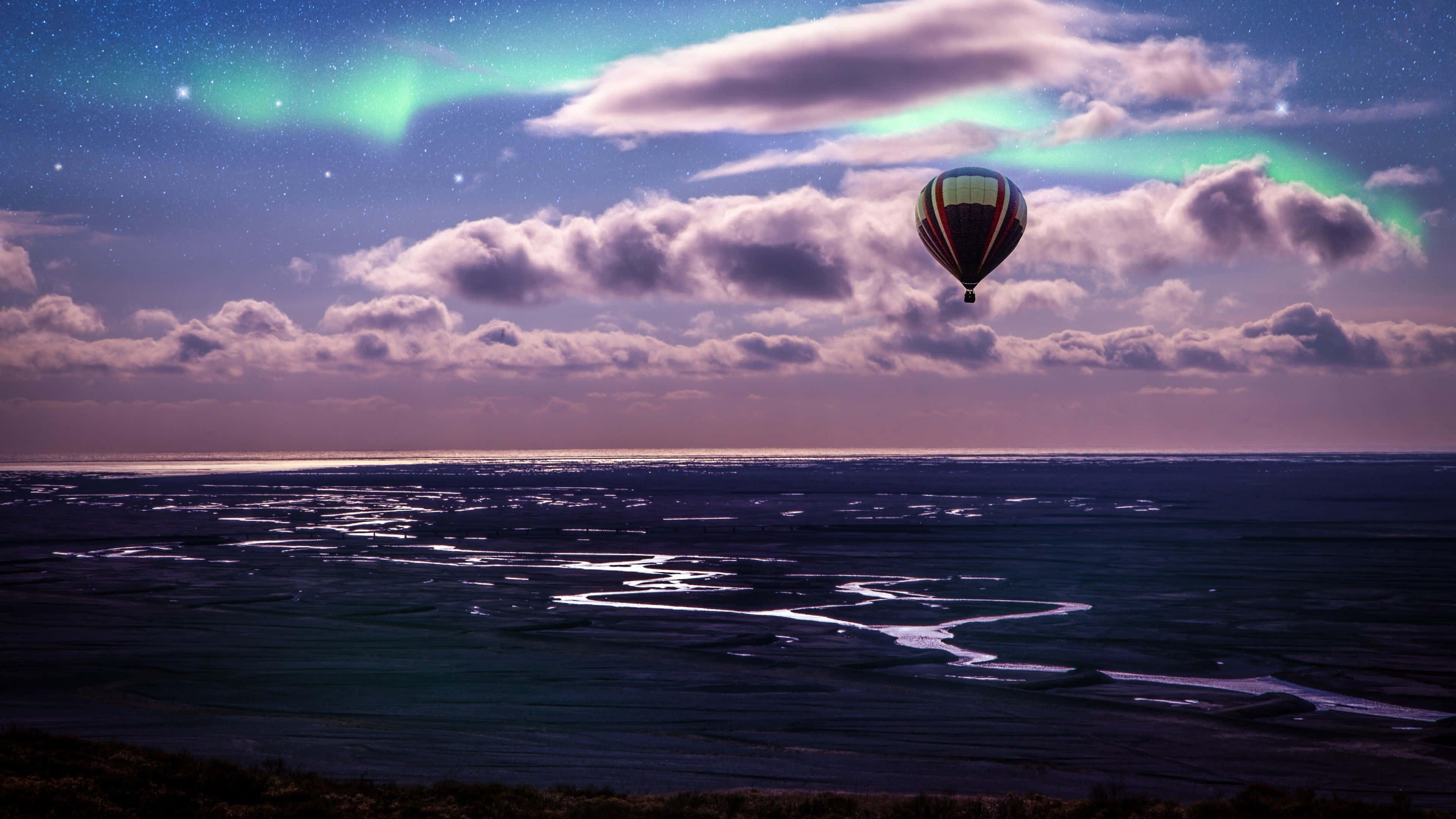 Hot Air Balloon 桌布 壁紙 — 一個熱氣球在繁星點點的夜空下，靜靜地漂浮在一條蜿蜒的河流之上，紫雲之上有充滿活力的綠色極光在舞動。