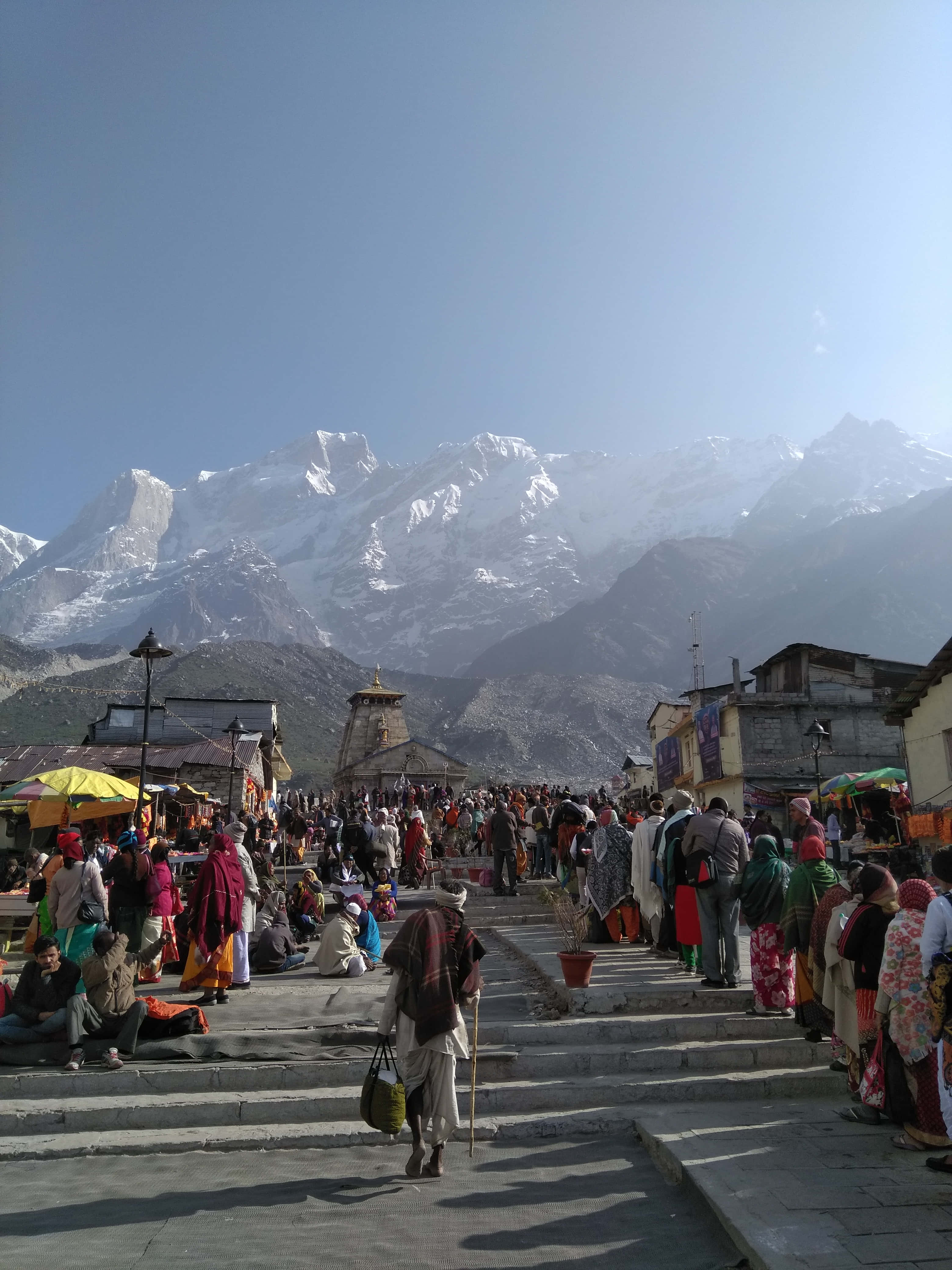Kedarnath 桌布 壁紙 — 一個熱鬧的山城，色彩繽紛的人群沿著石階向上攀登，朝向在晴朗藍天下雄偉的雪峰。
